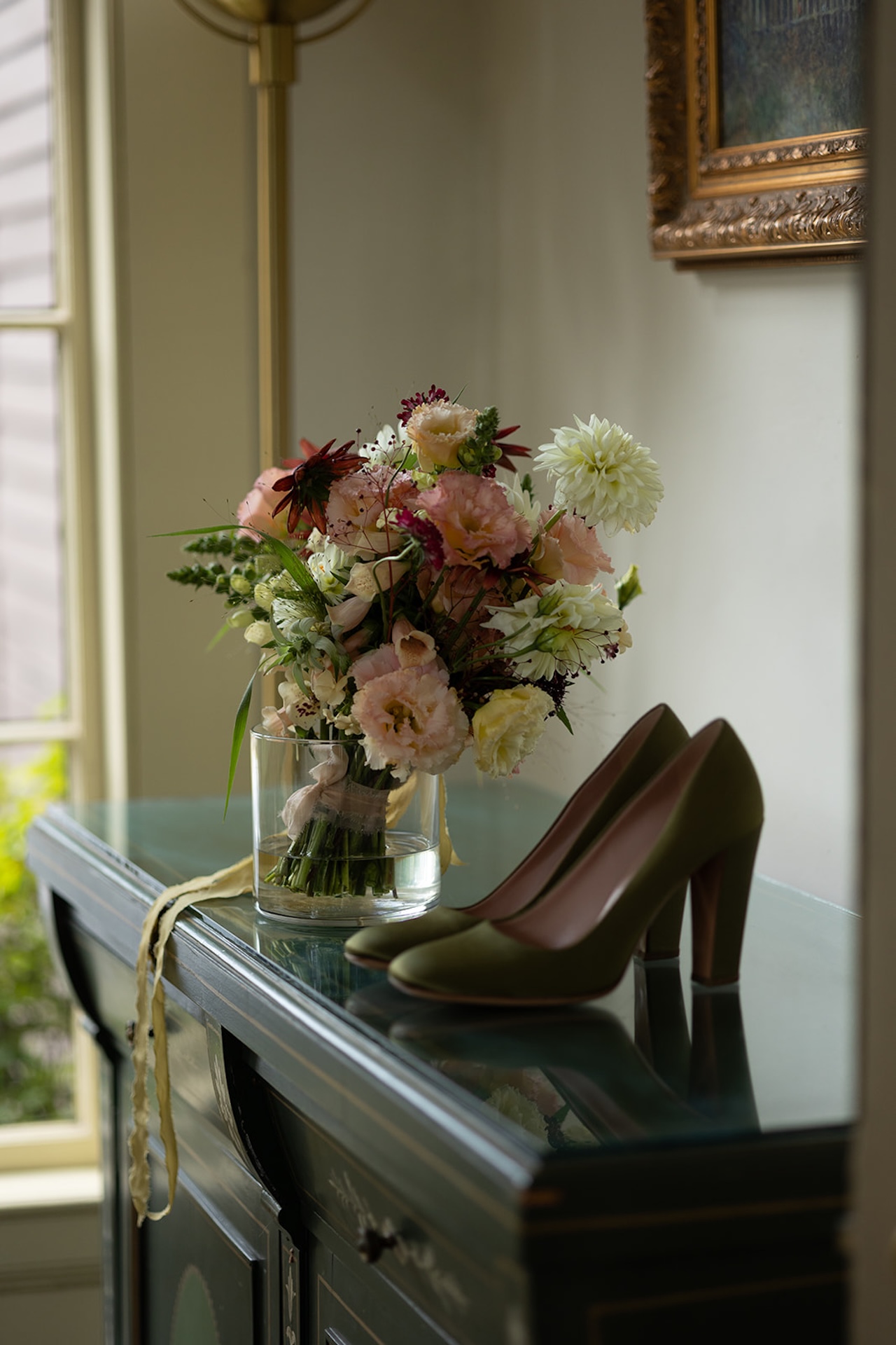 Bride’s bouquet and olive green heels displayed on a dark green antique cabinet, lit softly by a nearby window inside a Historic Wedding Venue.