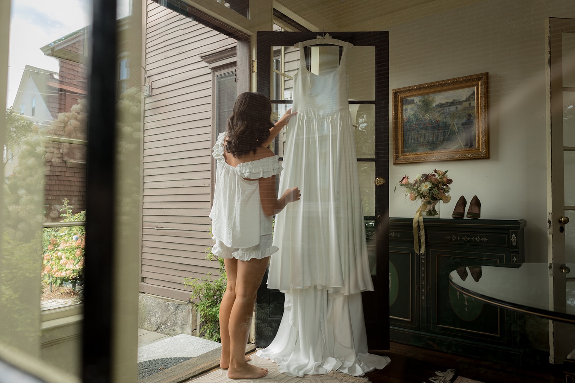 Bride looking at her wedding dress hanging on the glass door, captured through a reflective window for a layered, intimate feel.