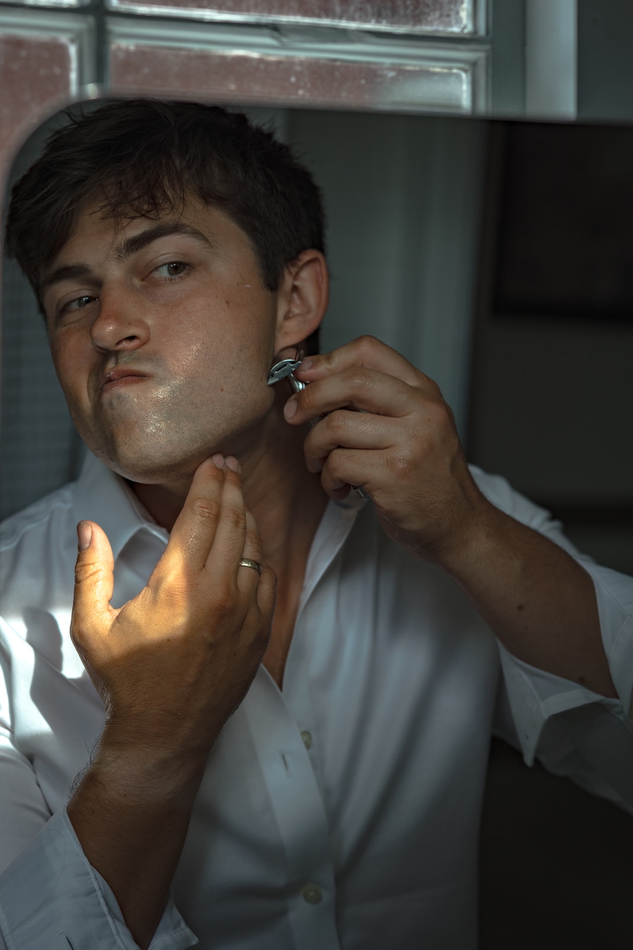 Groom shaving by the bathroom window, sunlight highlighting his face as he gets ready for the day.
