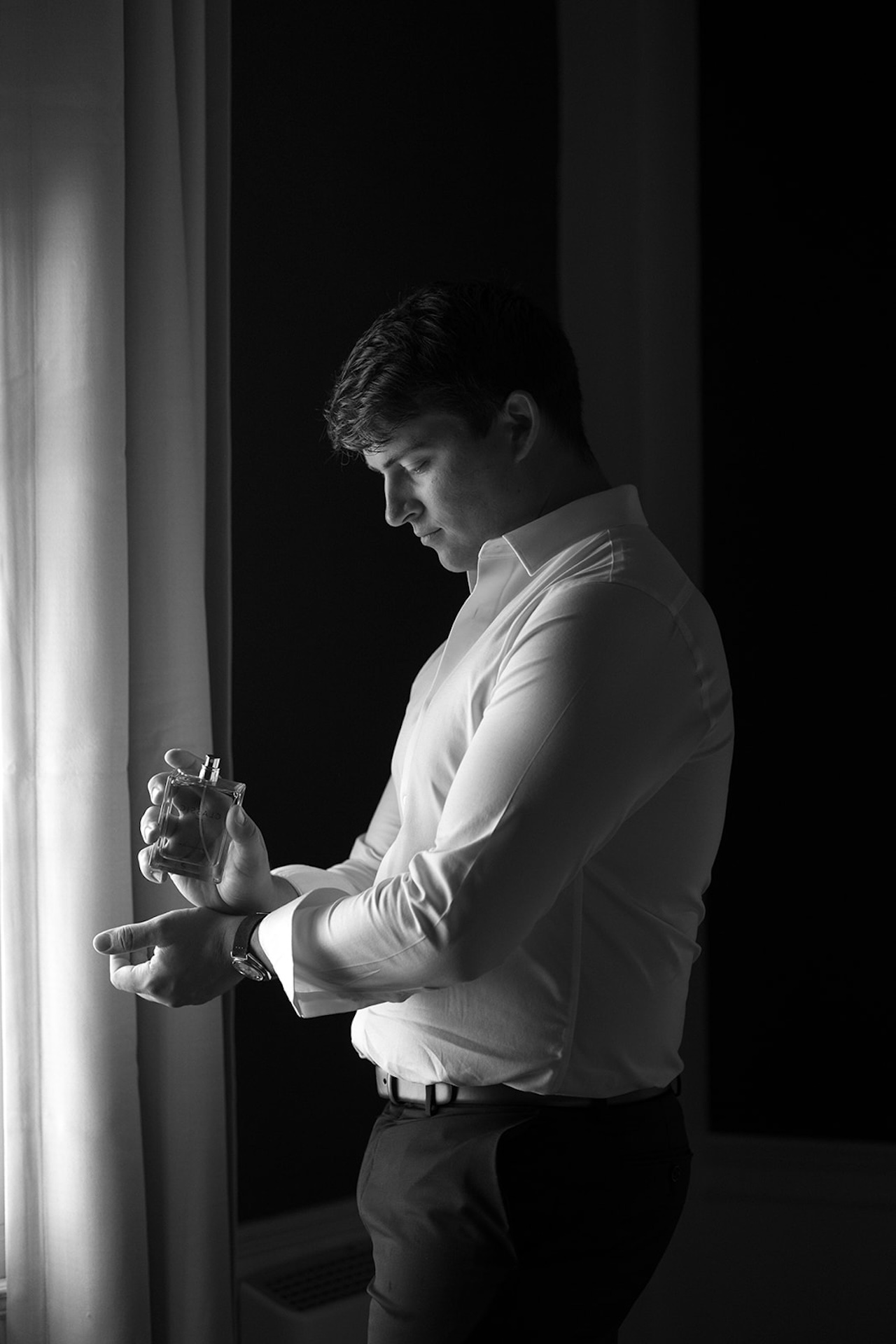Groom adjusting his cologne by a window, dramatic light highlighting his face and crisp white shirt in one of Maine’s Historic Wedding Venues.