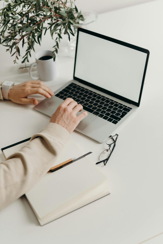 A detail photo of a person typing on their laptop with a notebook, pen, and glasses on the table.