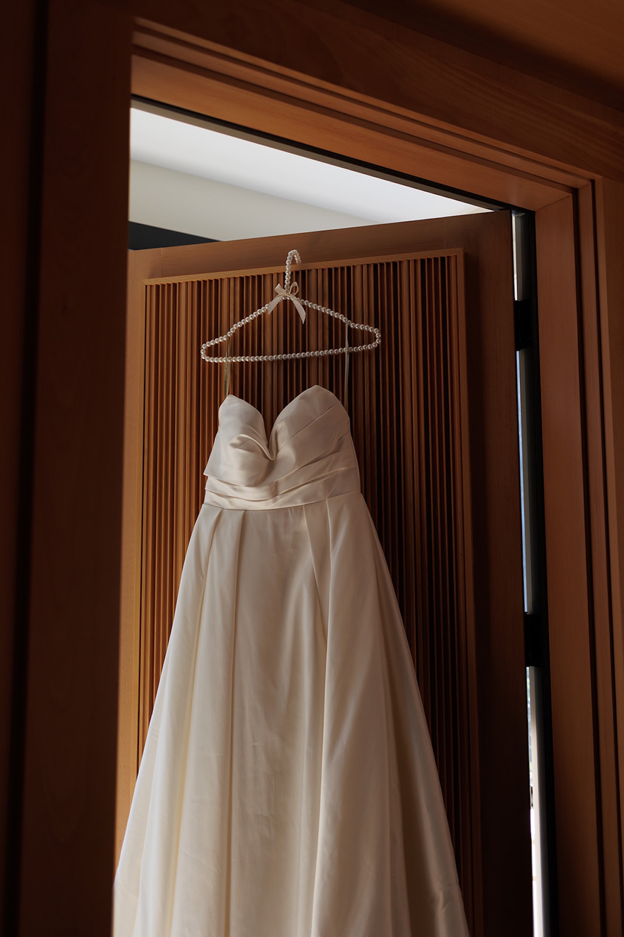 Strapless wedding gown hanging on a pearl hanger against a wooden door panel.
