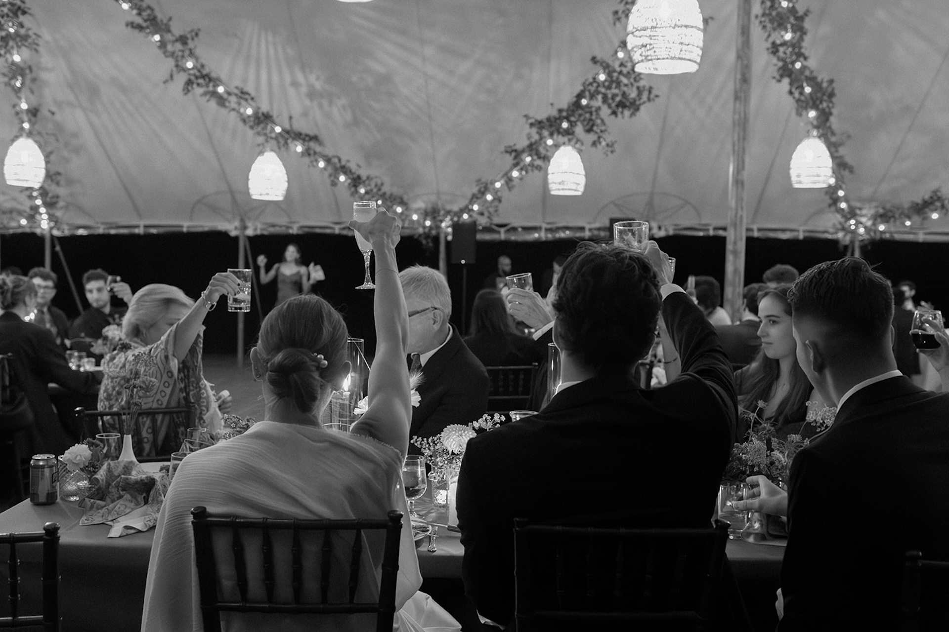 Guests raise glasses during a joyful reception toast under a tent filled with greenery and lights.