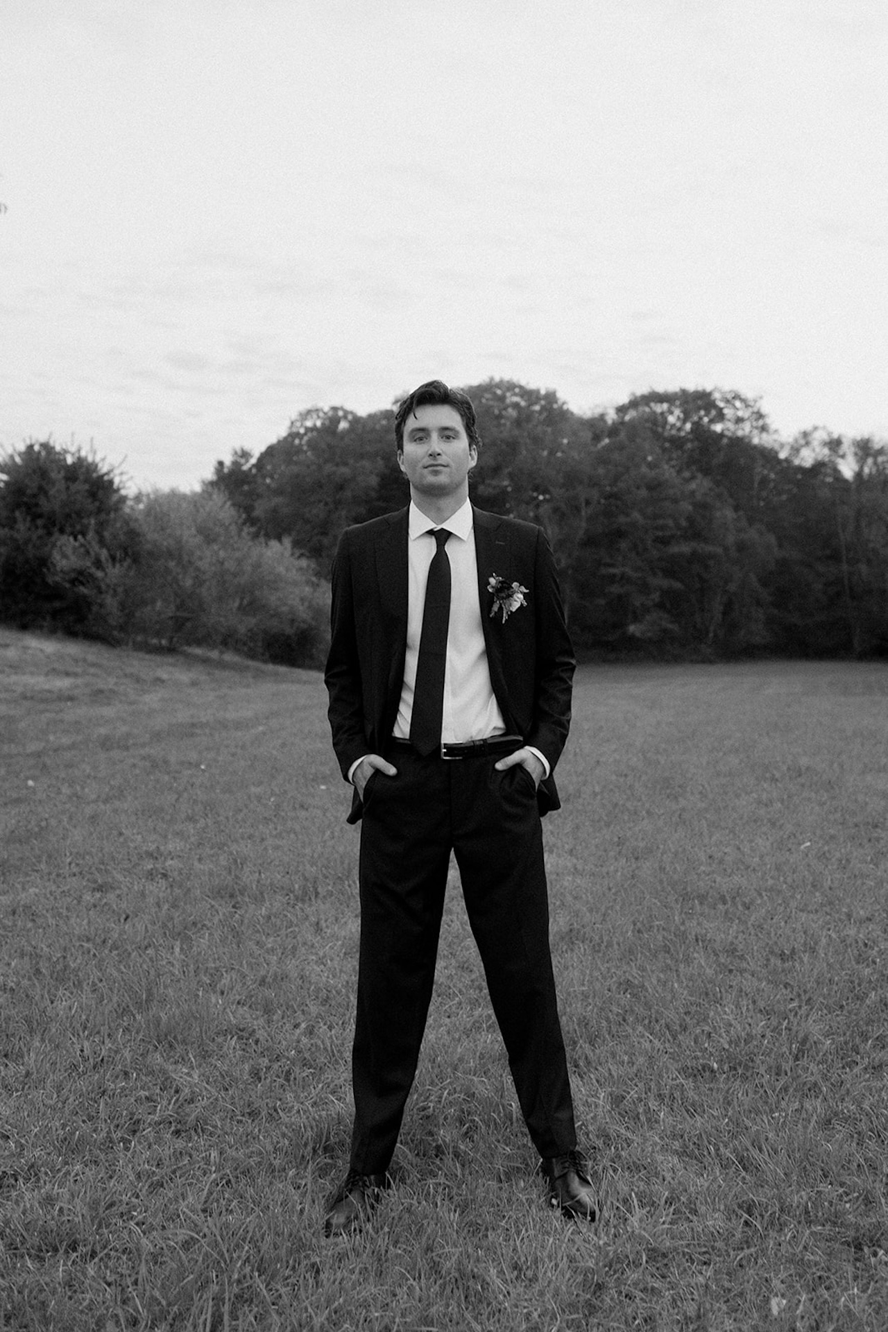 Groom stands confidently in an open field, hands in pockets, wearing a classic black suit.