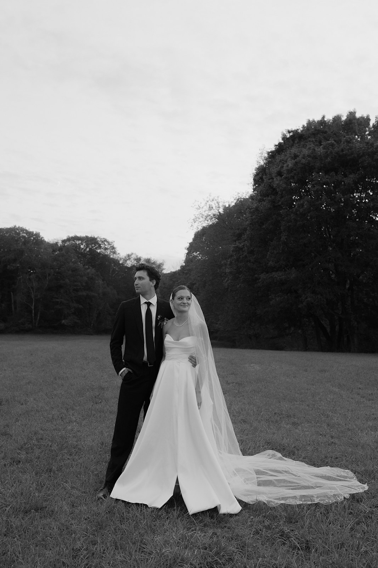 Black-and-white portrait of the bride and groom standing together in a meadow, looking off into the distance.