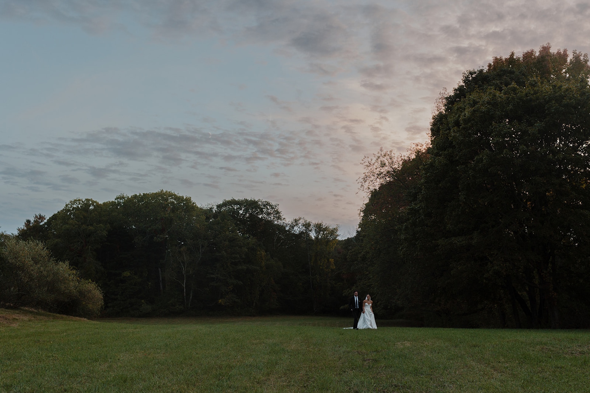 Couple walks across an open field at sunset during their Wedding in Maine.