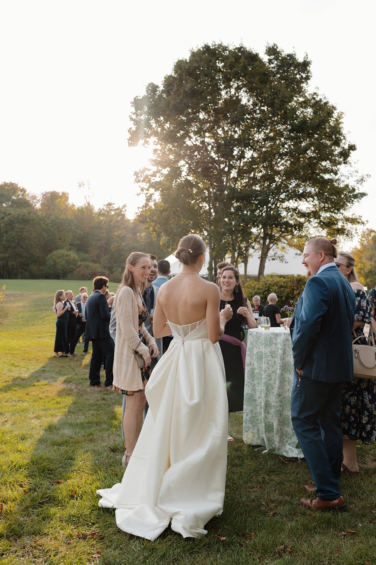 The bride chats with guests outdoors during cocktail hour, sunlight glowing behind her.