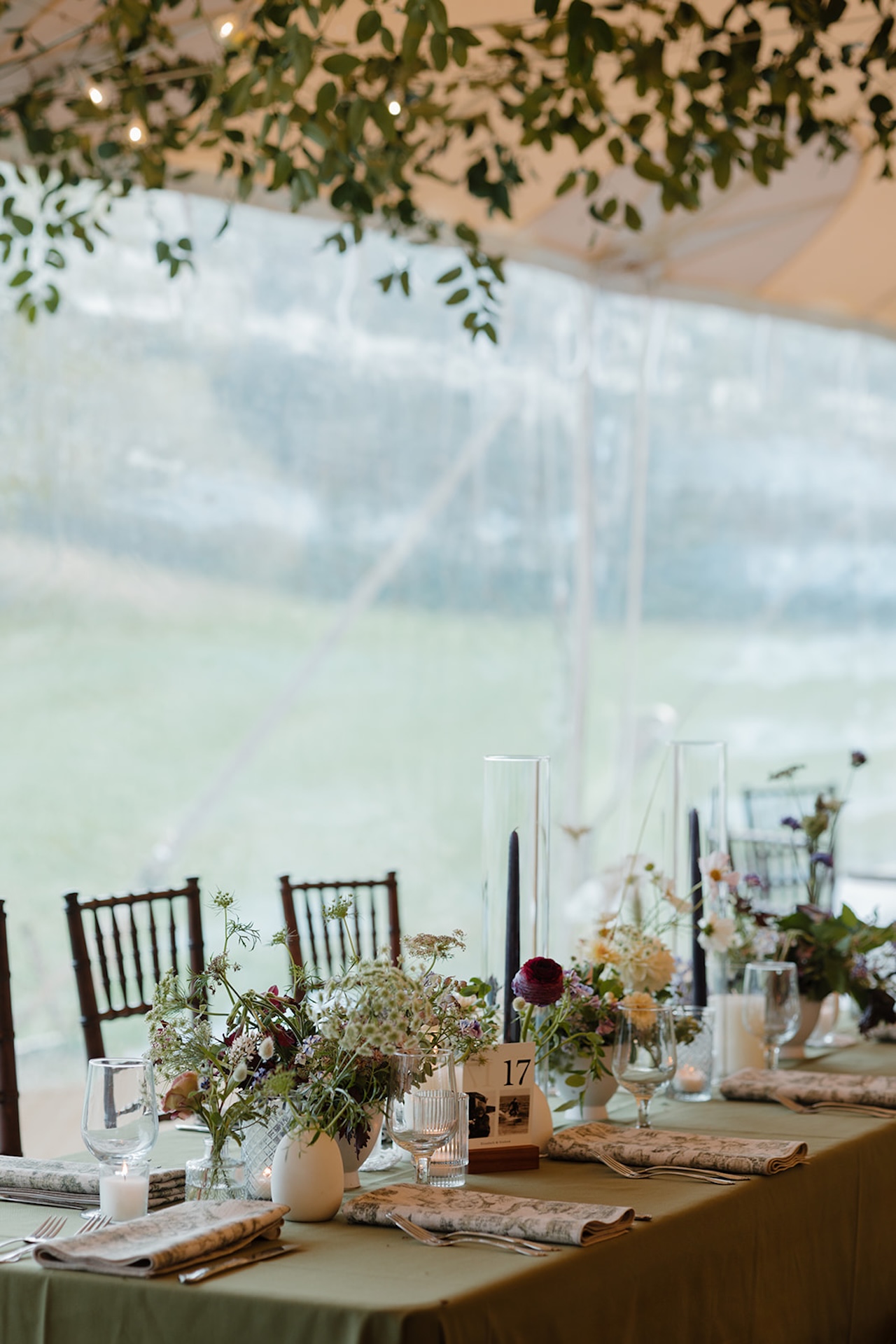 Romantic reception table under greenery with candles, florals, and folded napkins arranged for guests.