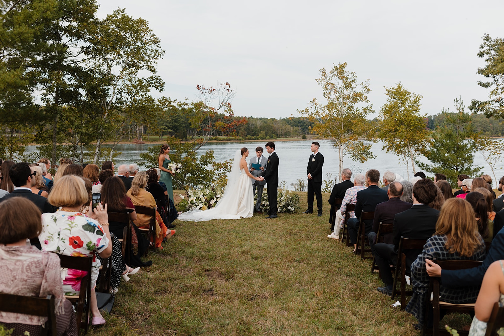 Lakeside ceremony scene as the couple exchanges vows surrounded by guests at their Wedding in Maine.
