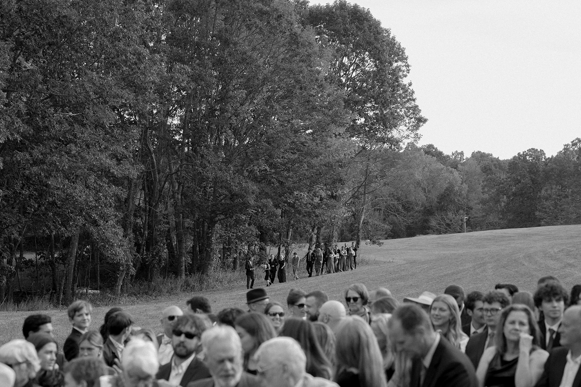 A black and white photo of all the guests in the foreground waiting for the ceremony to start as the wedding party, officiant, and everyone walks towards them.