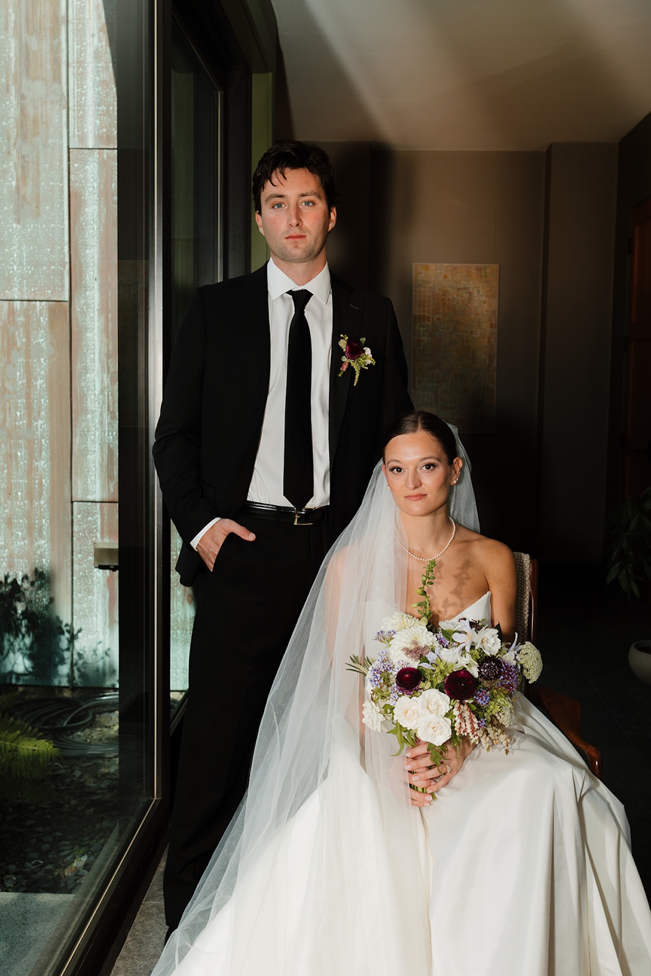 Modern editorial portrait of the bride and groom indoors, she seated with bouquet and he standing beside her.