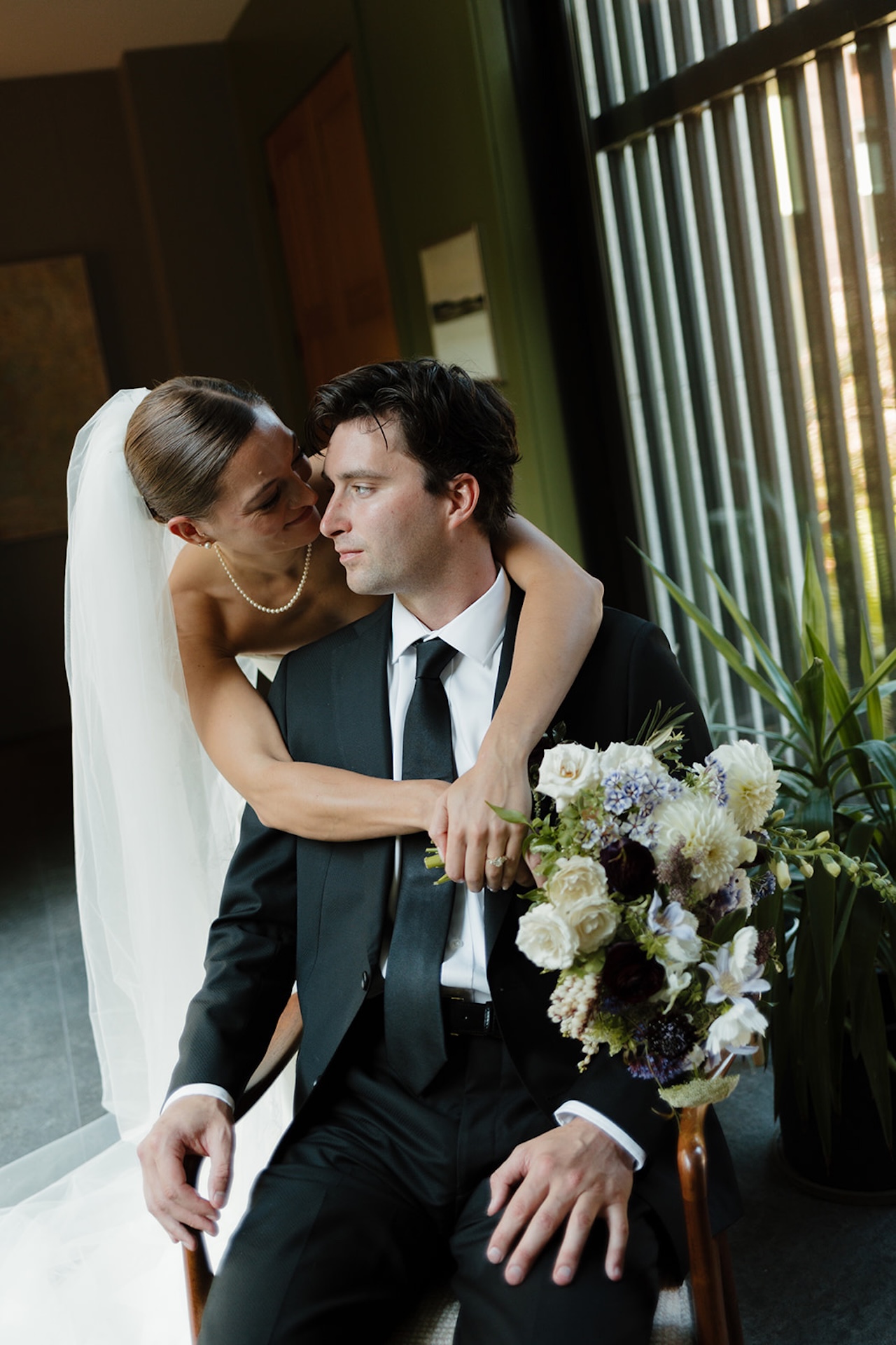 Bride wraps her arms around the groom while sitting indoors, sharing an intimate moment before portraits.