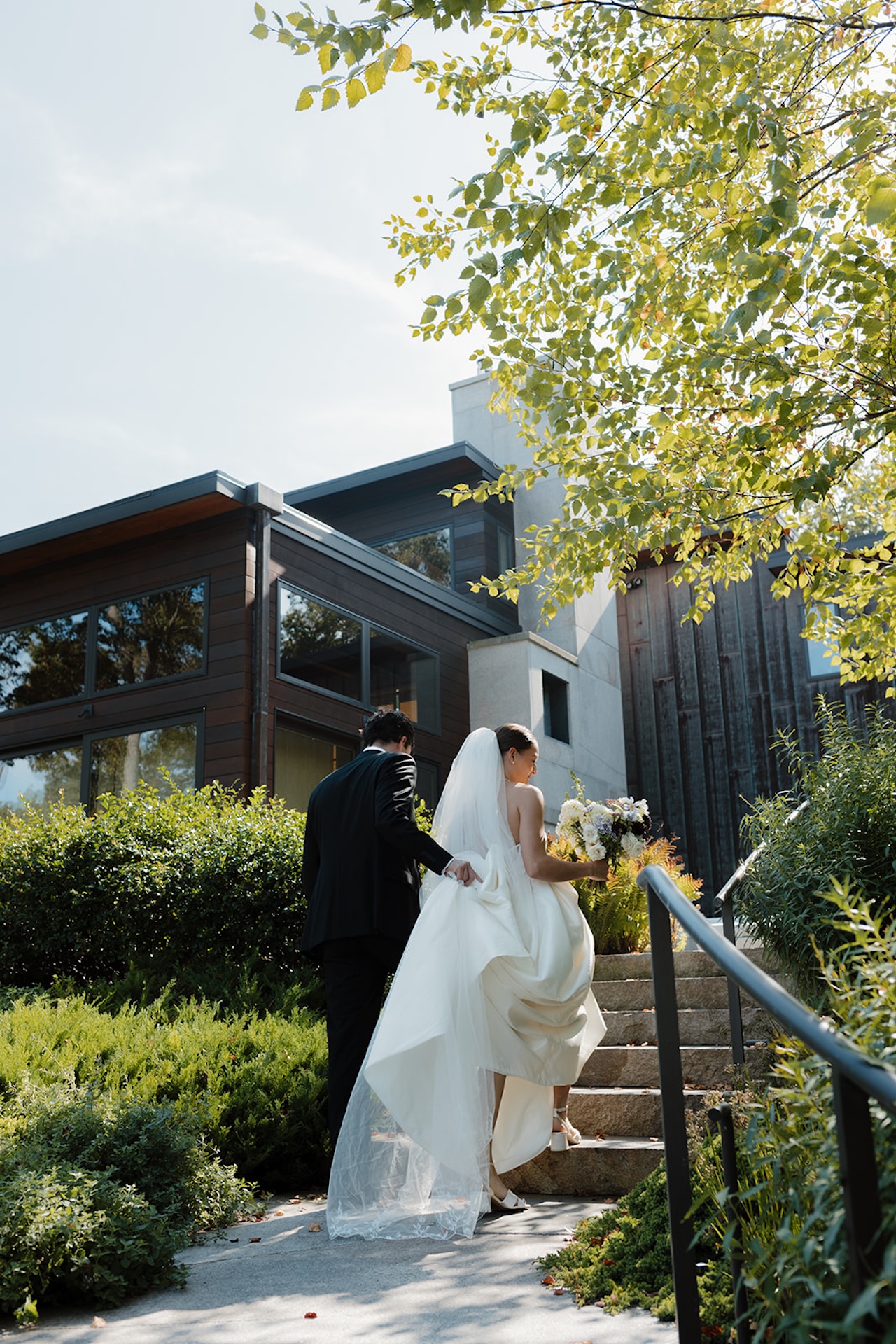 Bride and groom walk up stone steps outside a modern home surrounded by lush landscaping at their Wedding in Maine.