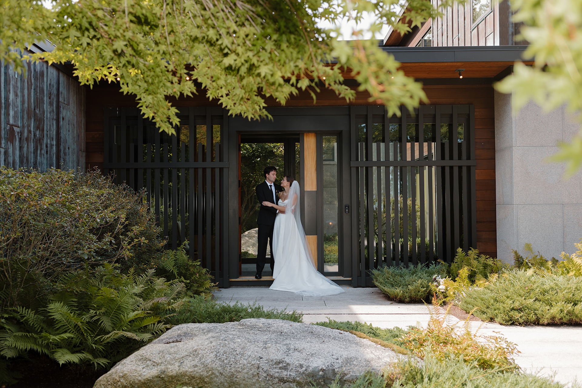 Couple stands framed in a modern entryway surrounded by greenery at their Wedding in Maine venue.