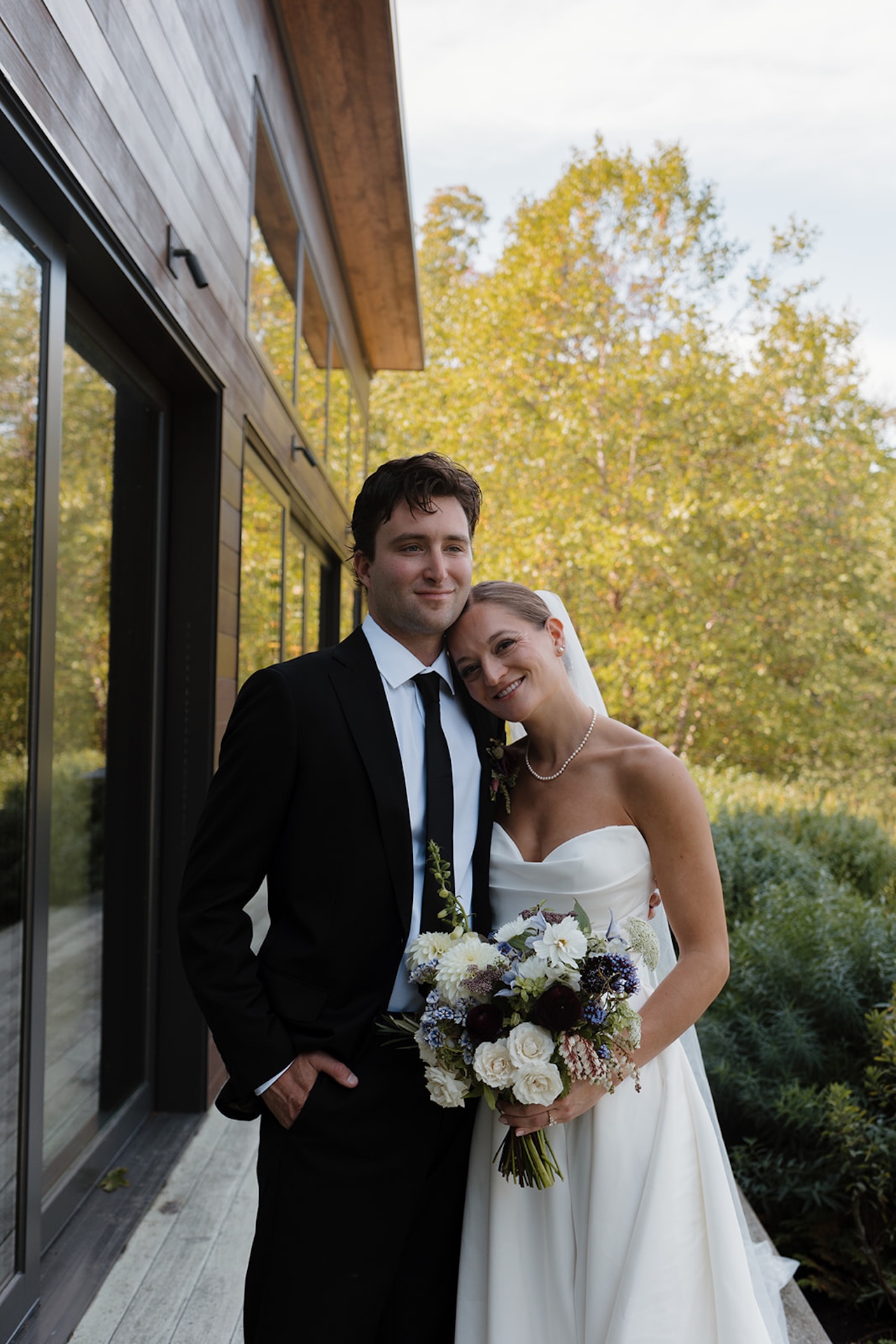 The couple stands close together outside, smiling as the bride rests her head on the groom’s shoulder.