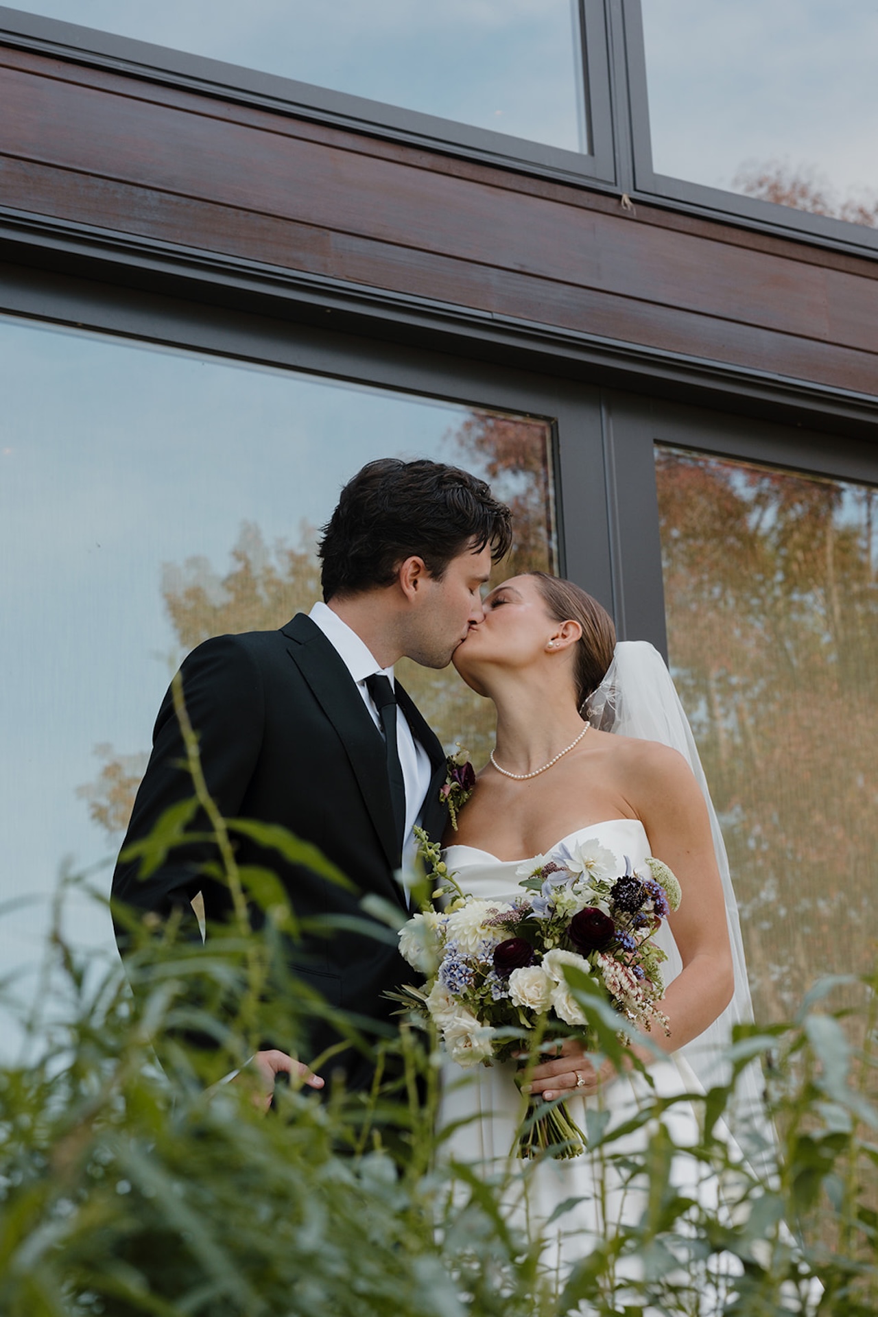Couple kisses outside a modern building surrounded by greenery during their Wedding in Maine.