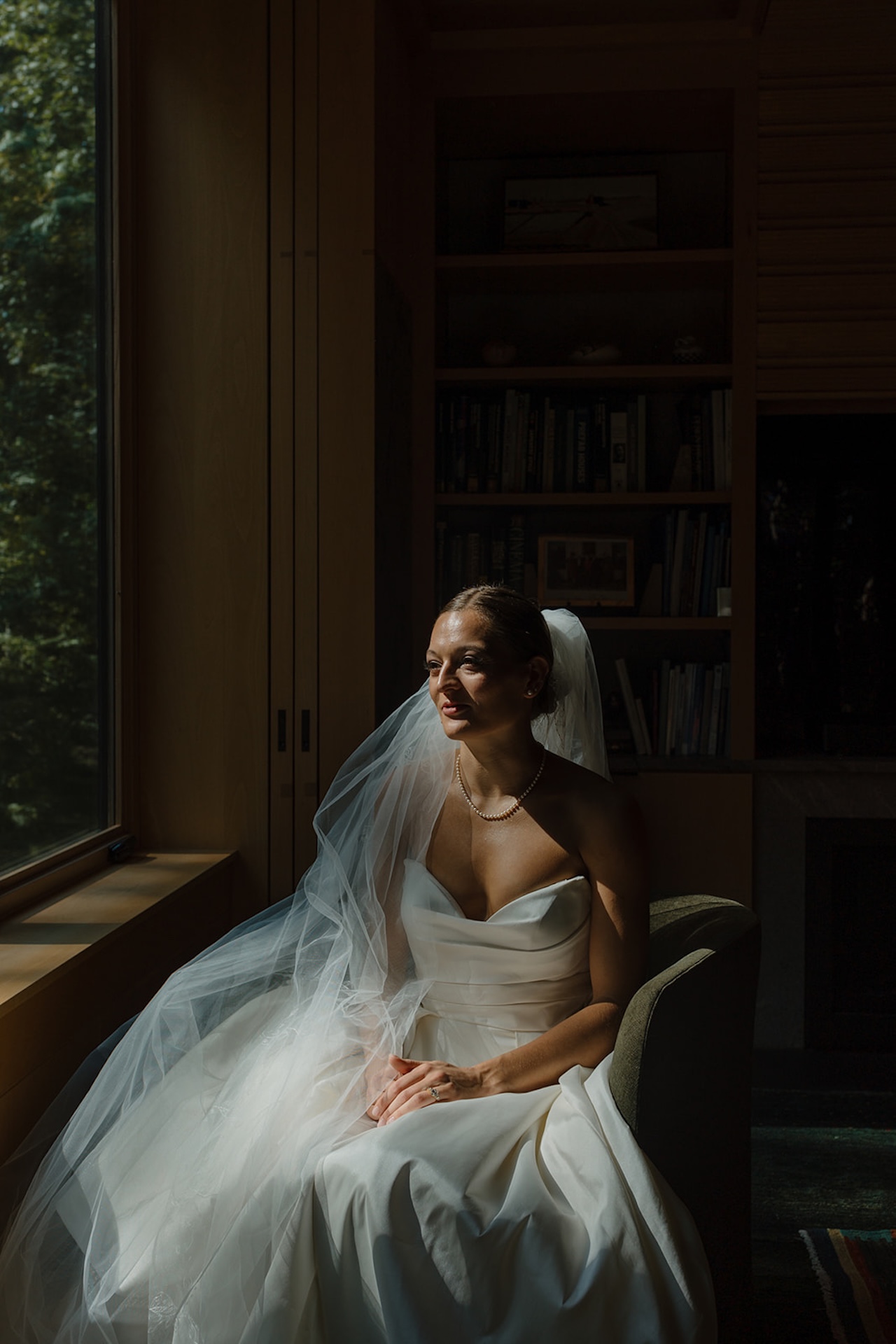 Bride sits alone in soft window light, veil draped around her shoulders at her Wedding in Maine.
