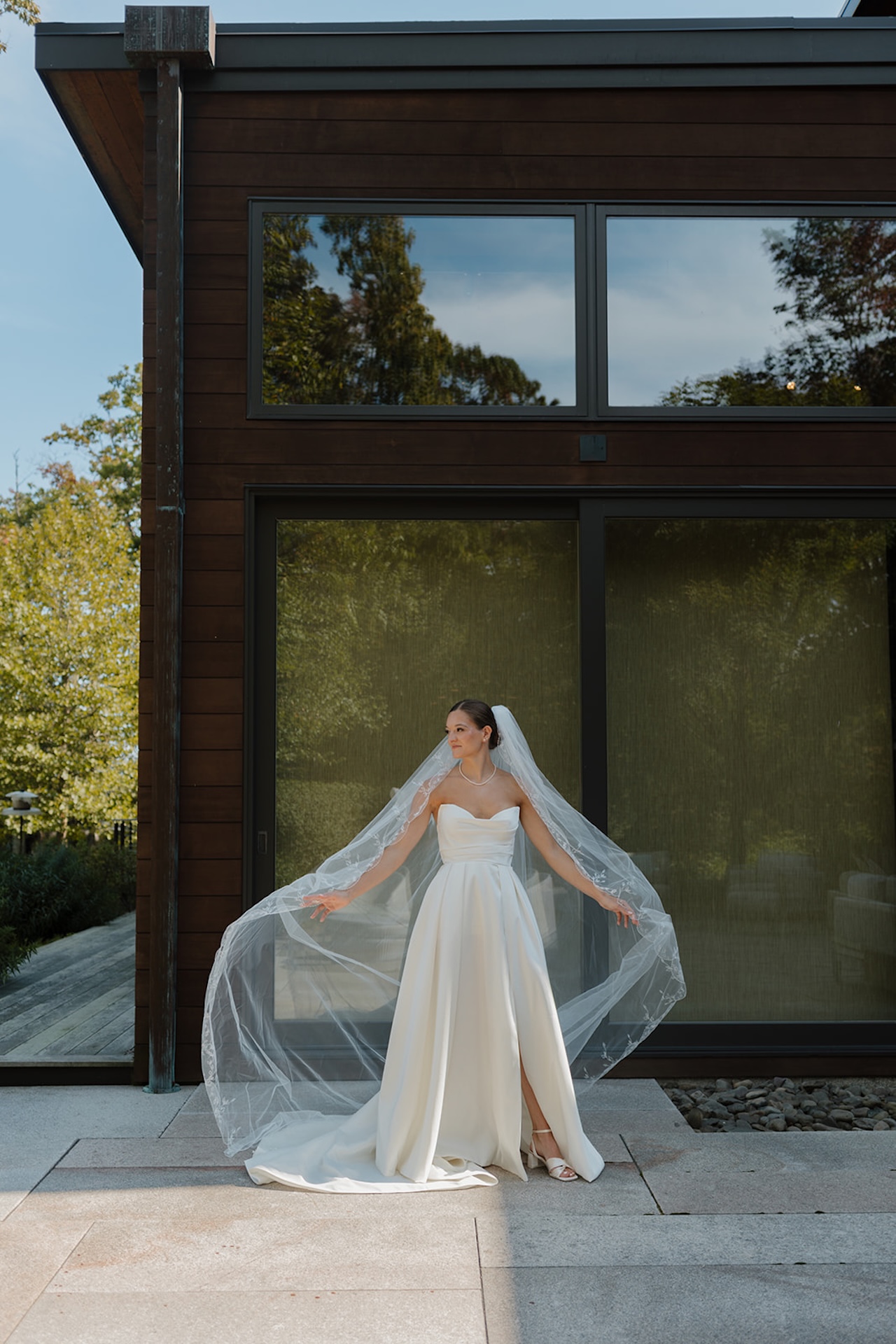 Bride stands outside holding her veil spread wide while wearing an elegant strapless gown.