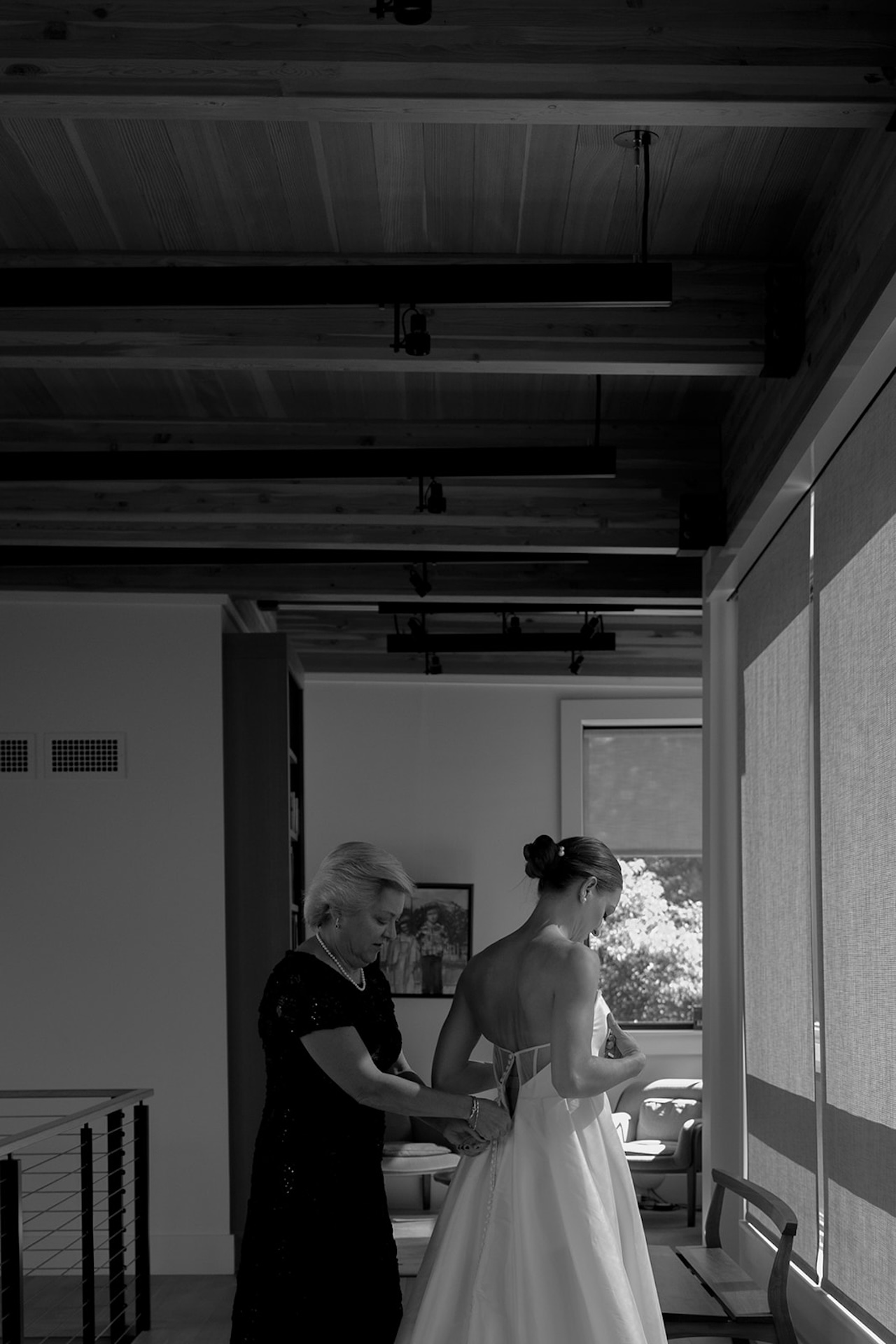 Mother buttons the bride’s gown as she stands by a sunlit window before the ceremony.