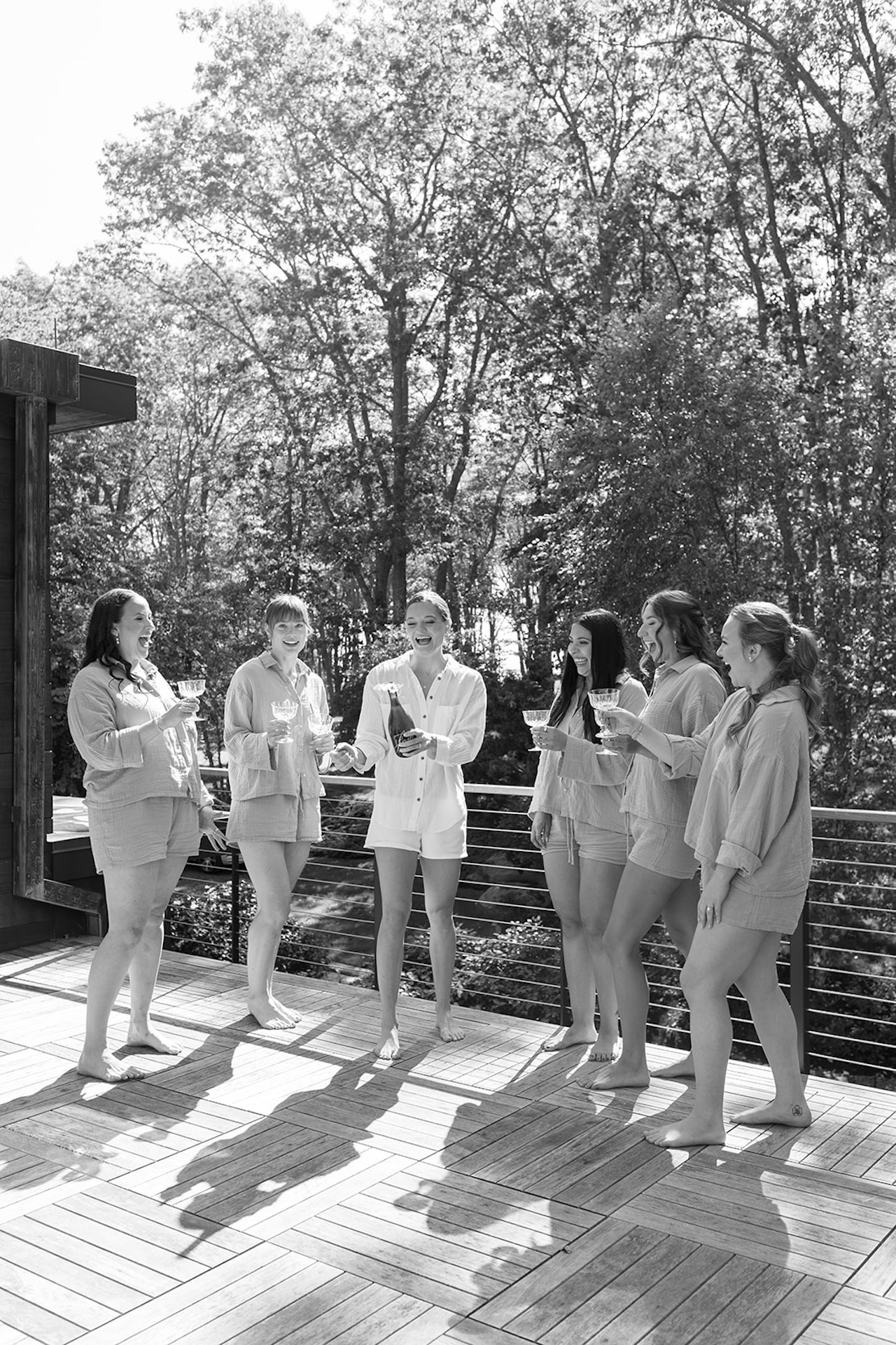 Bridesmaids and bride laugh together on a deck in matching loungewear while celebrating pre-ceremony.