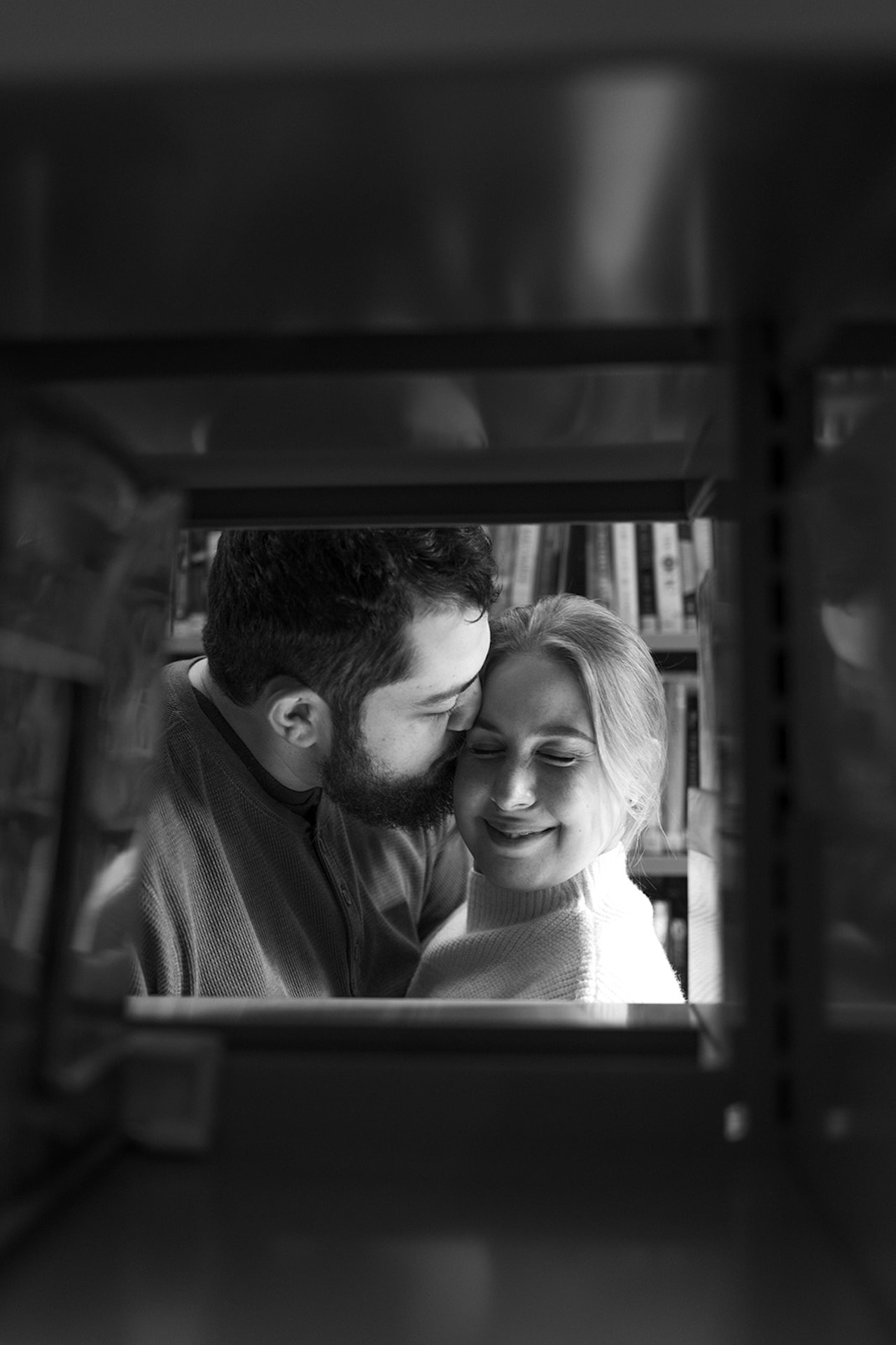 Black and white engagement photo of a couple sharing a quiet moment between bookshelves, highlighting timeless Engagement Photo Session Ideas with emotional depth.