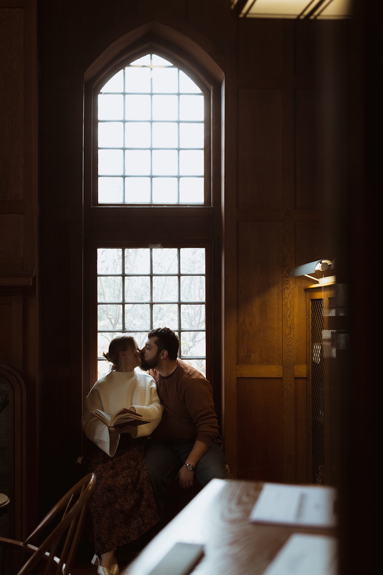 Library engagement photo of a couple sharing a quiet kiss by a tall window, highlighting creative Engagement Photo Session Ideas in a cozy indoor location.