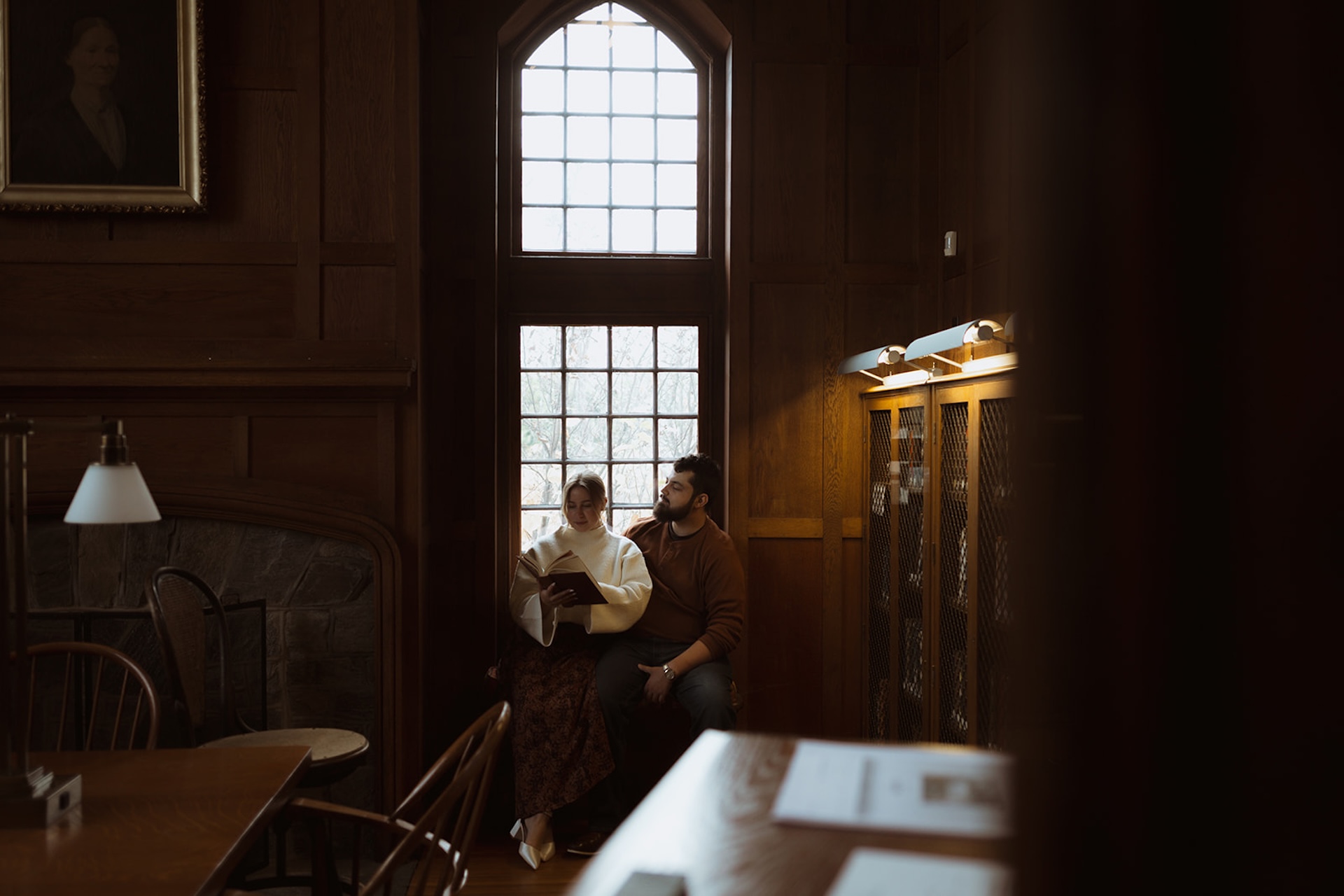 Engagement portrait of a couple seated by a tall window reading together, surrounded by wood-paneled walls and soft natural light.