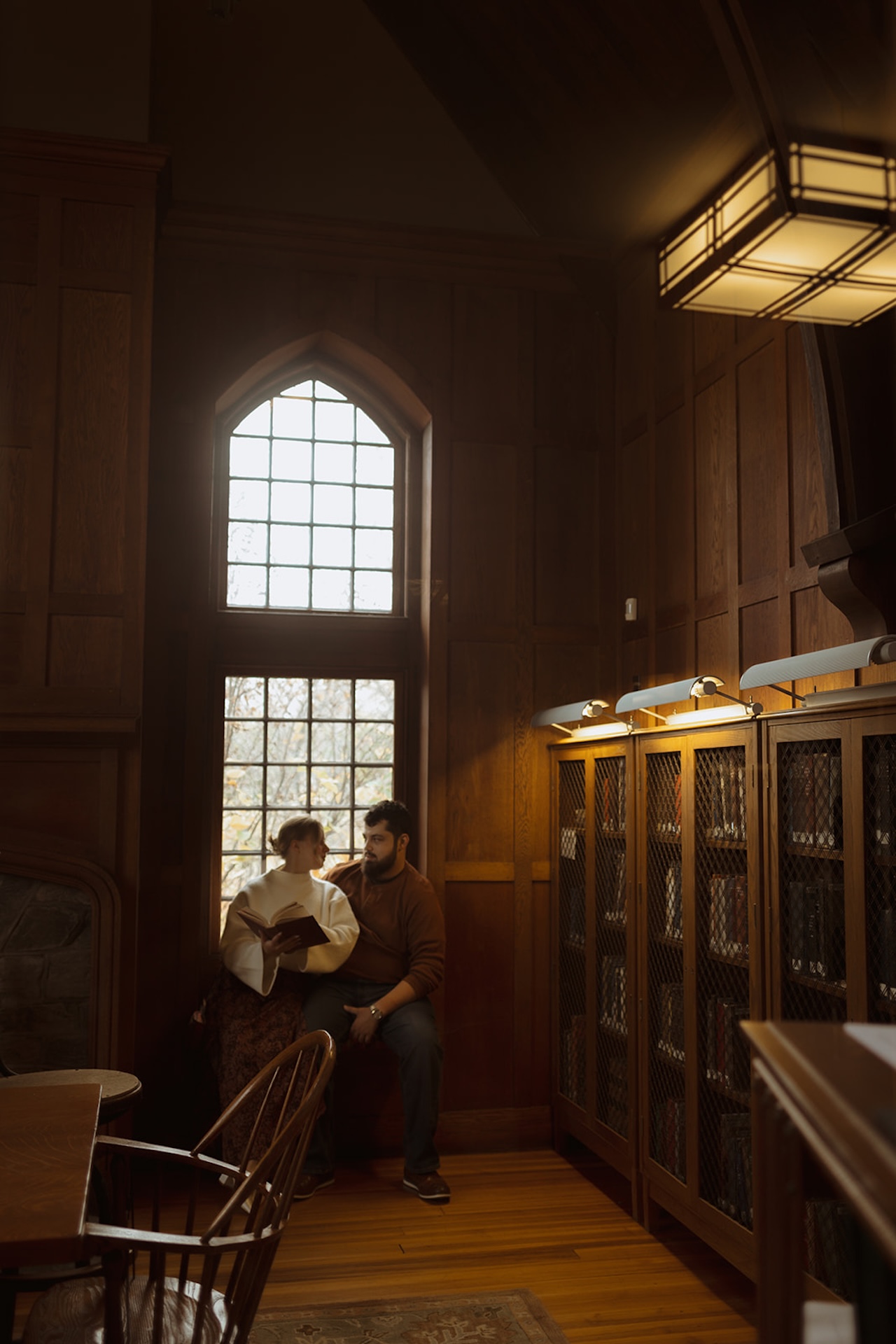 Wide engagement photo of a couple sitting together beneath tall arched windows in a historic library, highlighting classic Engagement Photo Session Ideas with natural light.