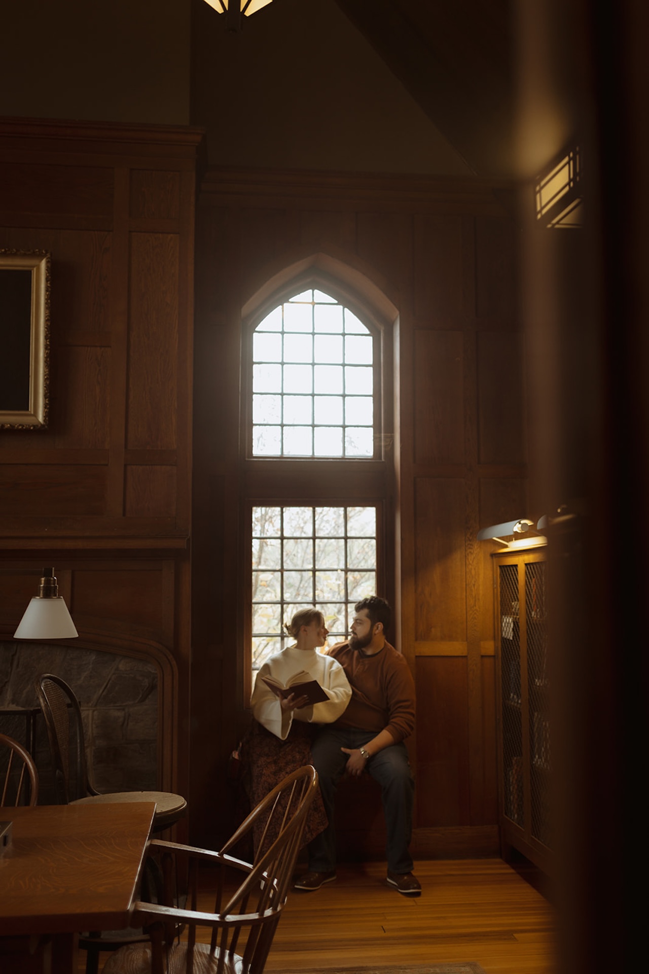 Wide engagement photo of a couple sitting together beneath tall arched windows in a historic library, highlighting classic Engagement Photo Session Ideas with natural light.