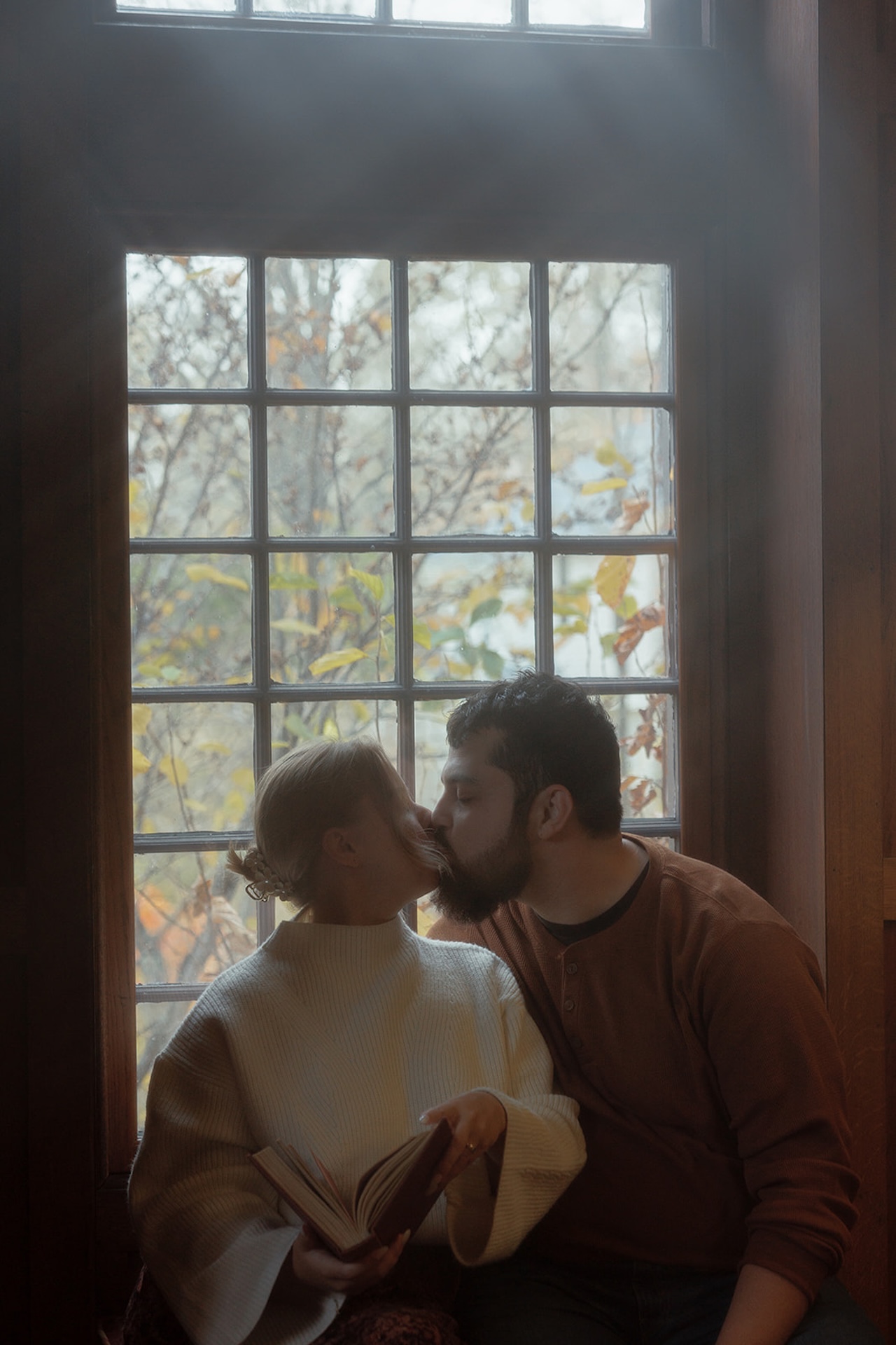 Romantic engagement photo of a couple kissing by a window while holding a book, creating a warm and intimate indoor scene.