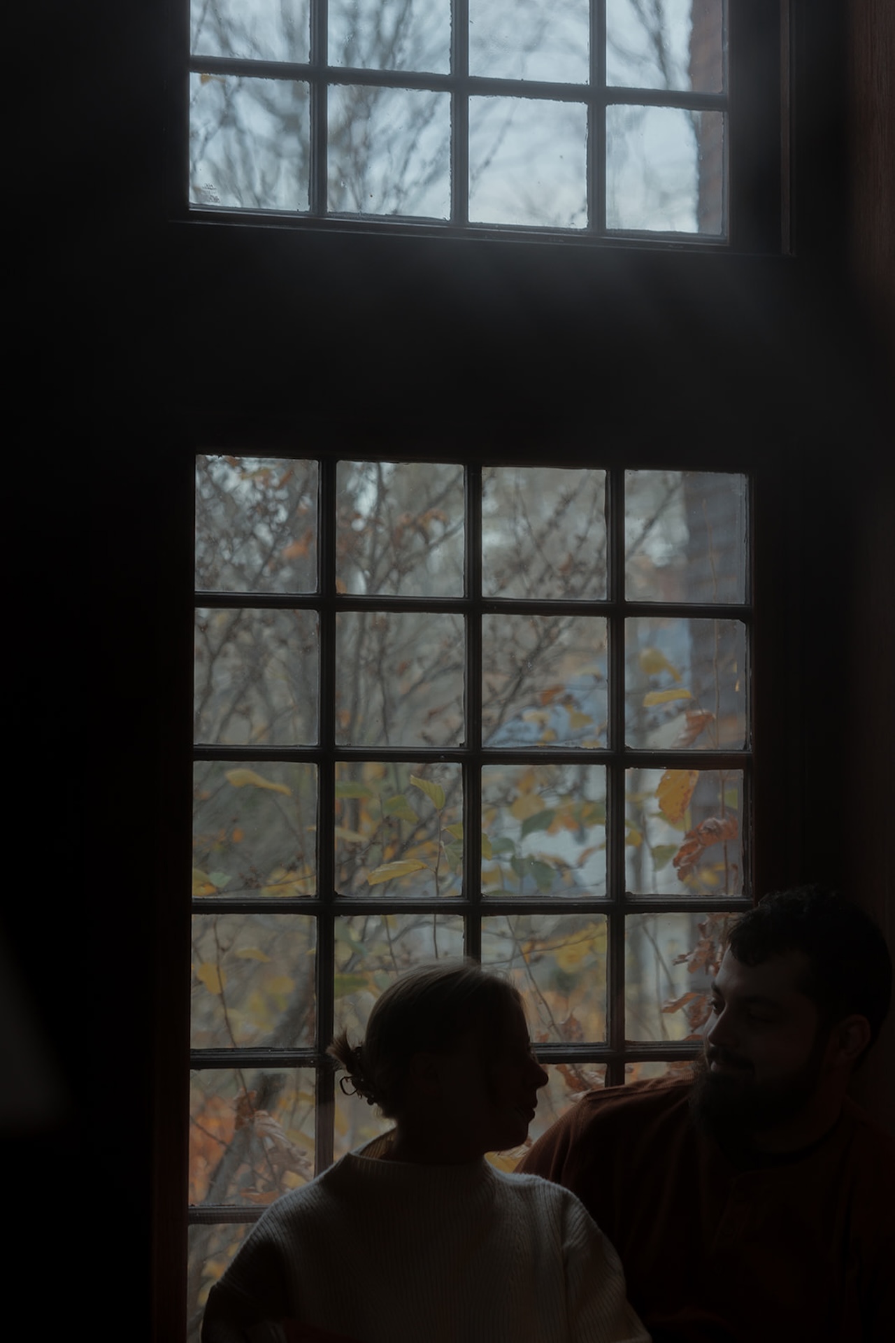 Romantic engagement photo of a couple chatting in front of a large window with autumn trees outside, showcasing cozy Engagement Photo Session Ideas indoors.