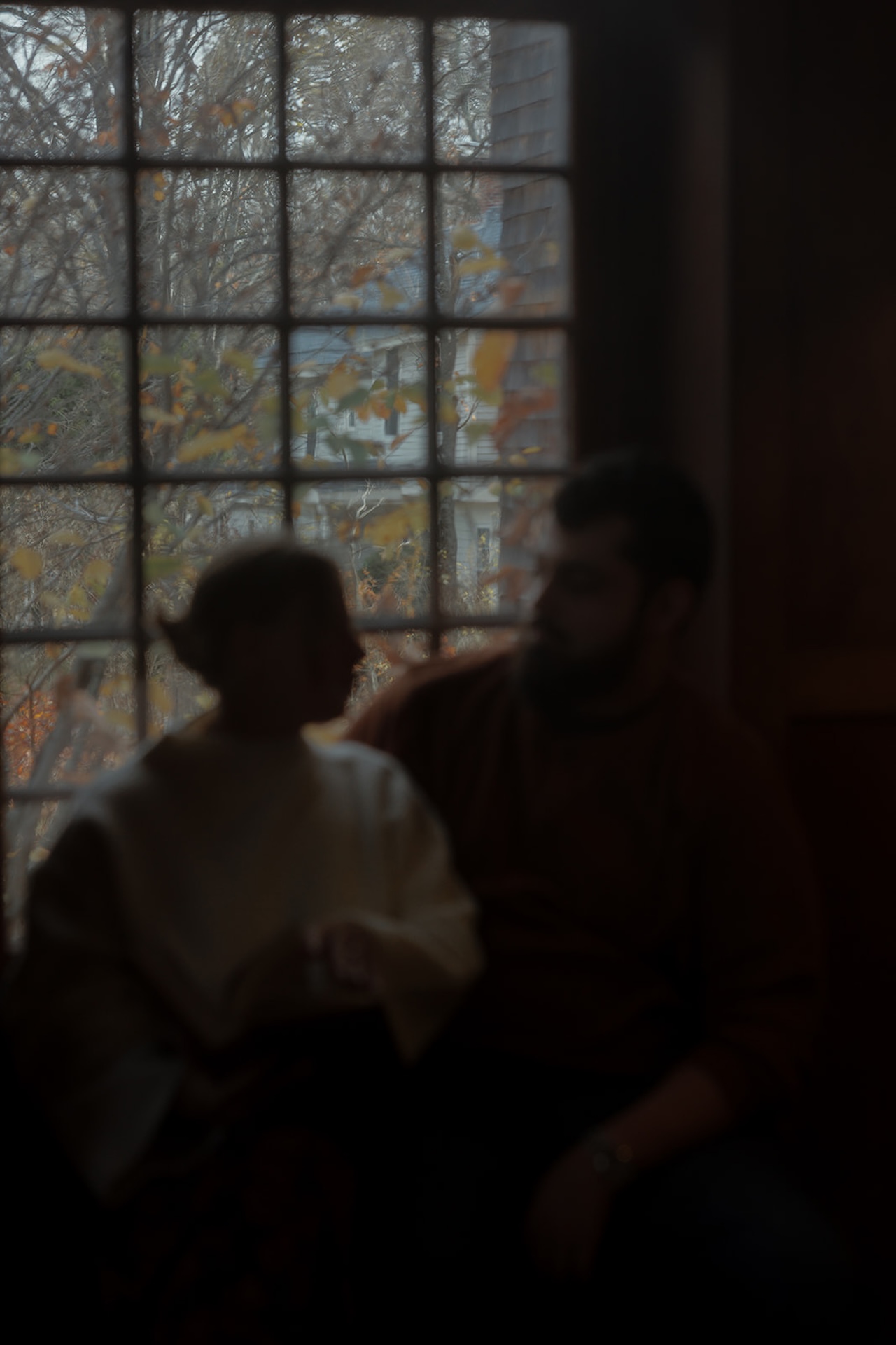 Soft-focus engagement photo of a couple sitting together by a large window, framed by autumn leaves outside for a quiet, cinematic moment.