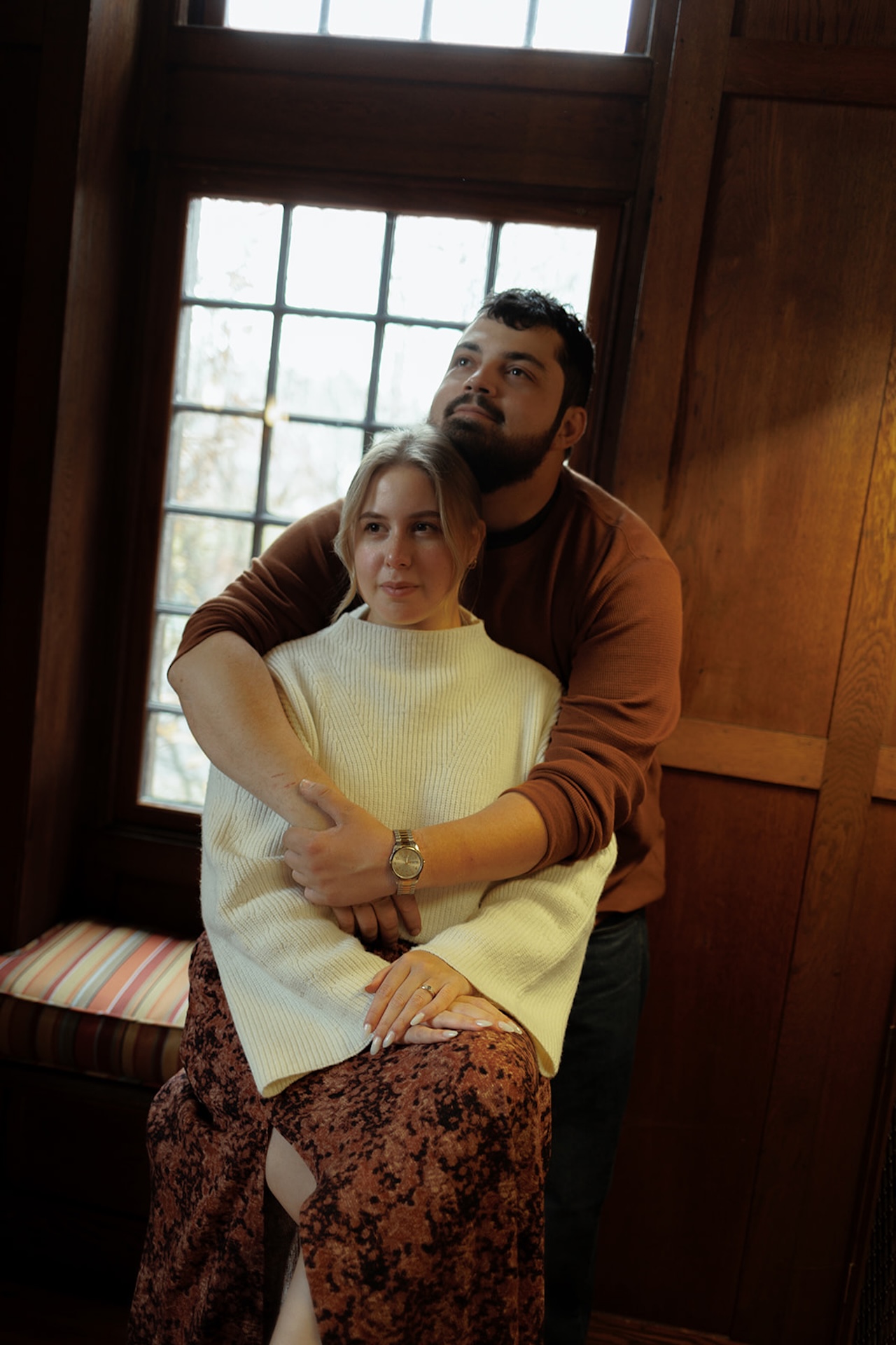 Cozy indoor engagement portrait of a couple by a window, wrapped in warm layers and natural light, creating a relaxed and intimate feel.