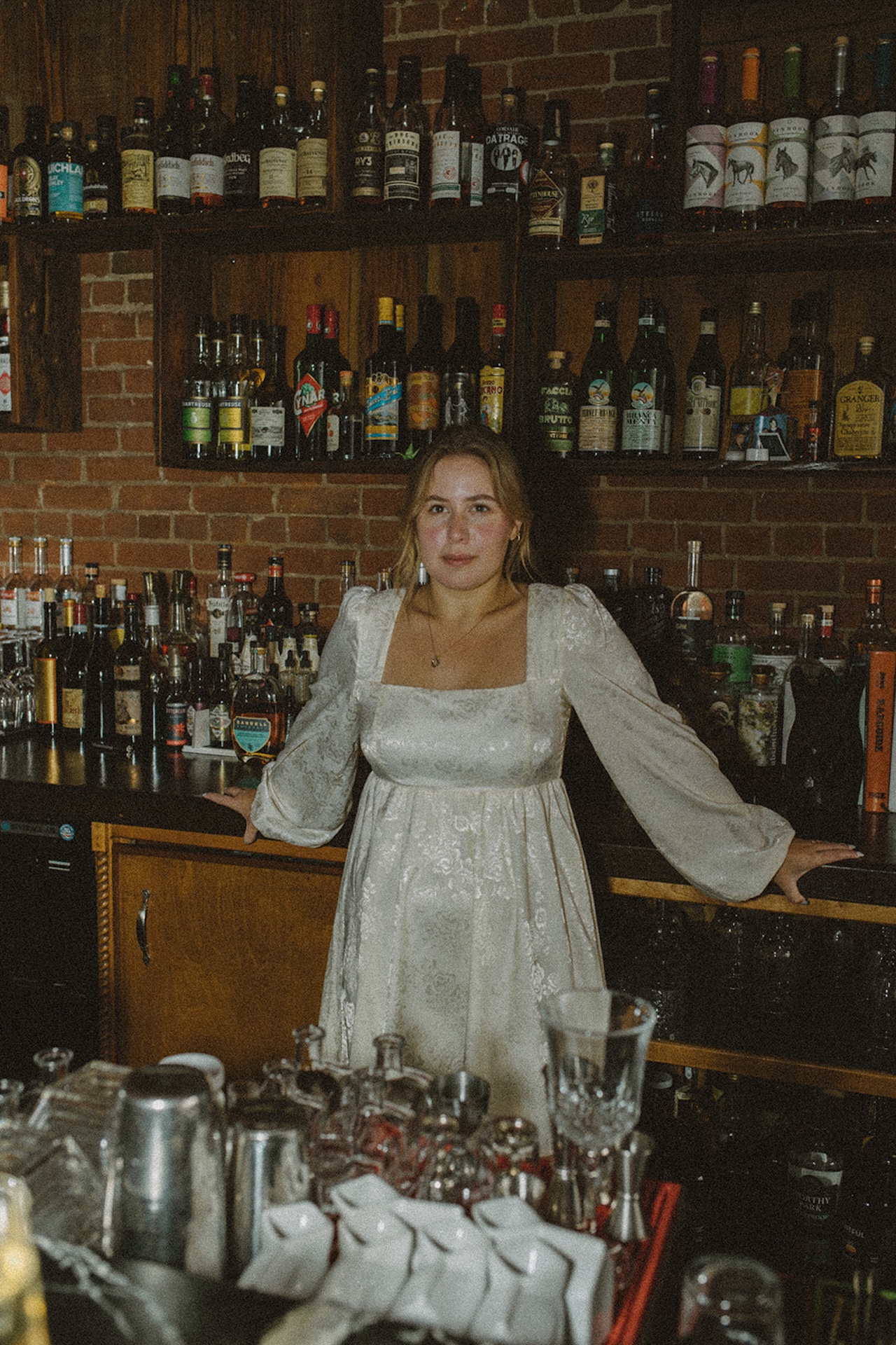 Editorial engagement portrait of a woman standing confidently behind a bar in a flowing white dress, showcasing bold Engagement Photo Session Ideas with personality.