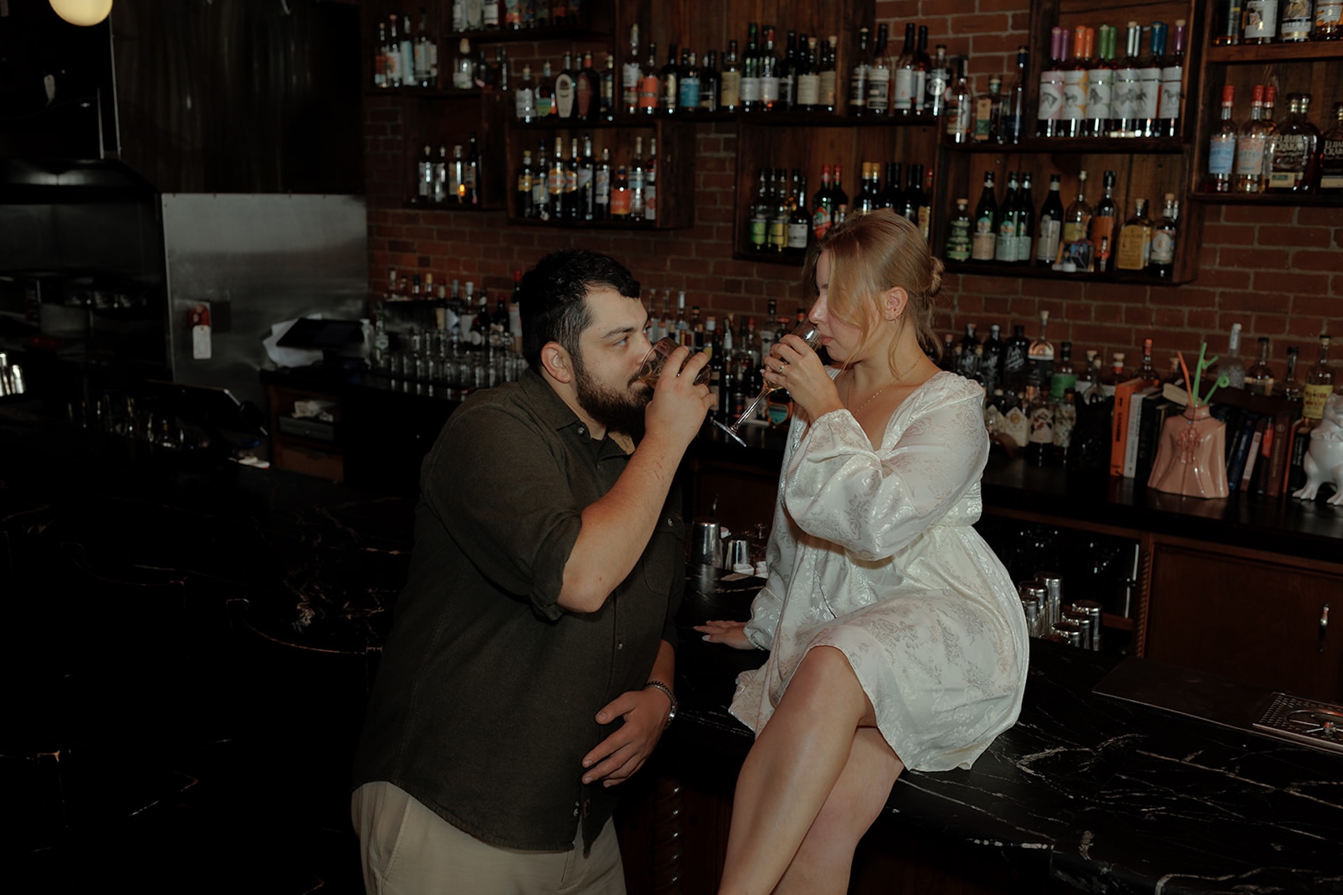 Playful engagement photo of a couple sipping drinks together at a bar, sitting close and laughing in a warm, intimate setting.