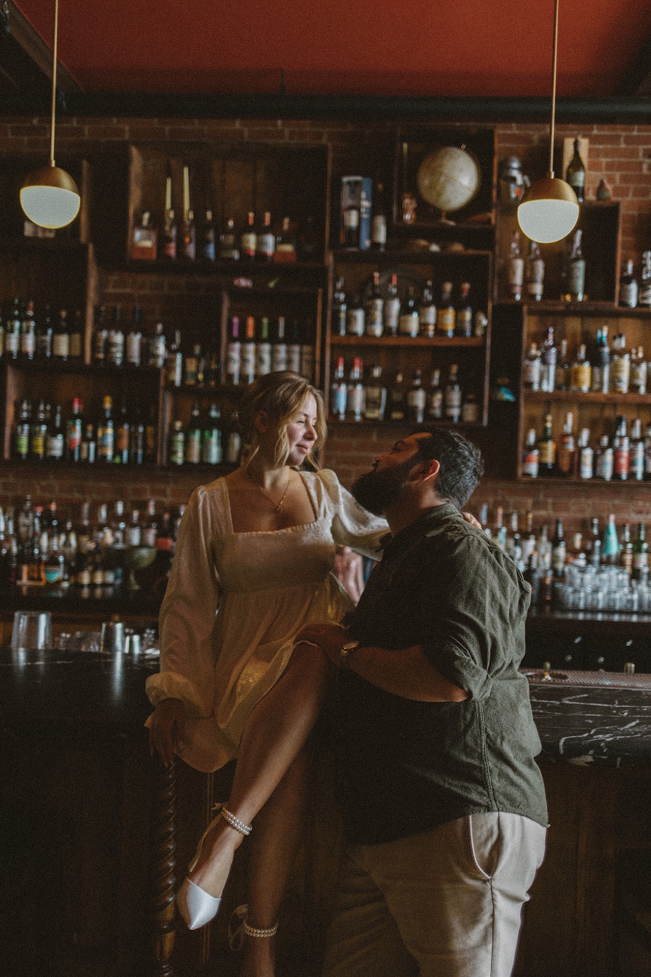 Editorial-style engagement photo of a couple leaning together at a bar surrounded by shelves of bottles, showcasing unique Engagement Photo Session Ideas with cinematic vibes.