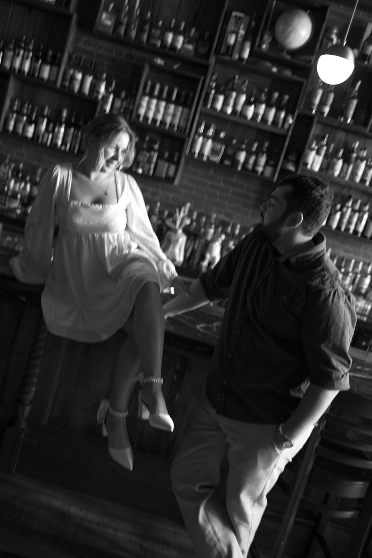Black and white engagement photo of a woman sitting on a bar counter laughing while her partner stands beside her, showing playful Engagement Photo Session Ideas indoors.