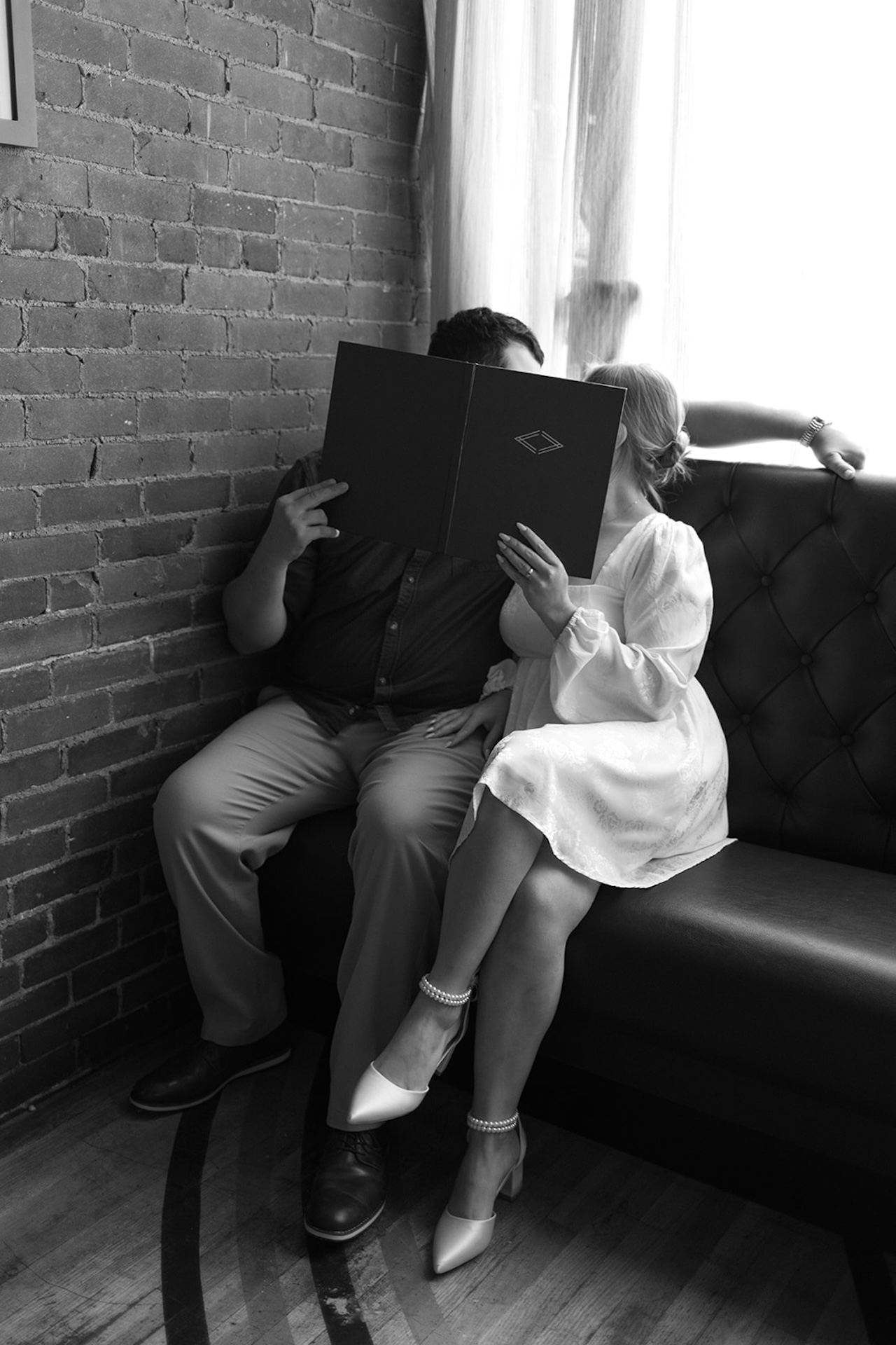 Black and white engagement photo of a couple sitting close together on a couch, sharing a quiet moment behind a book near a brick wall.