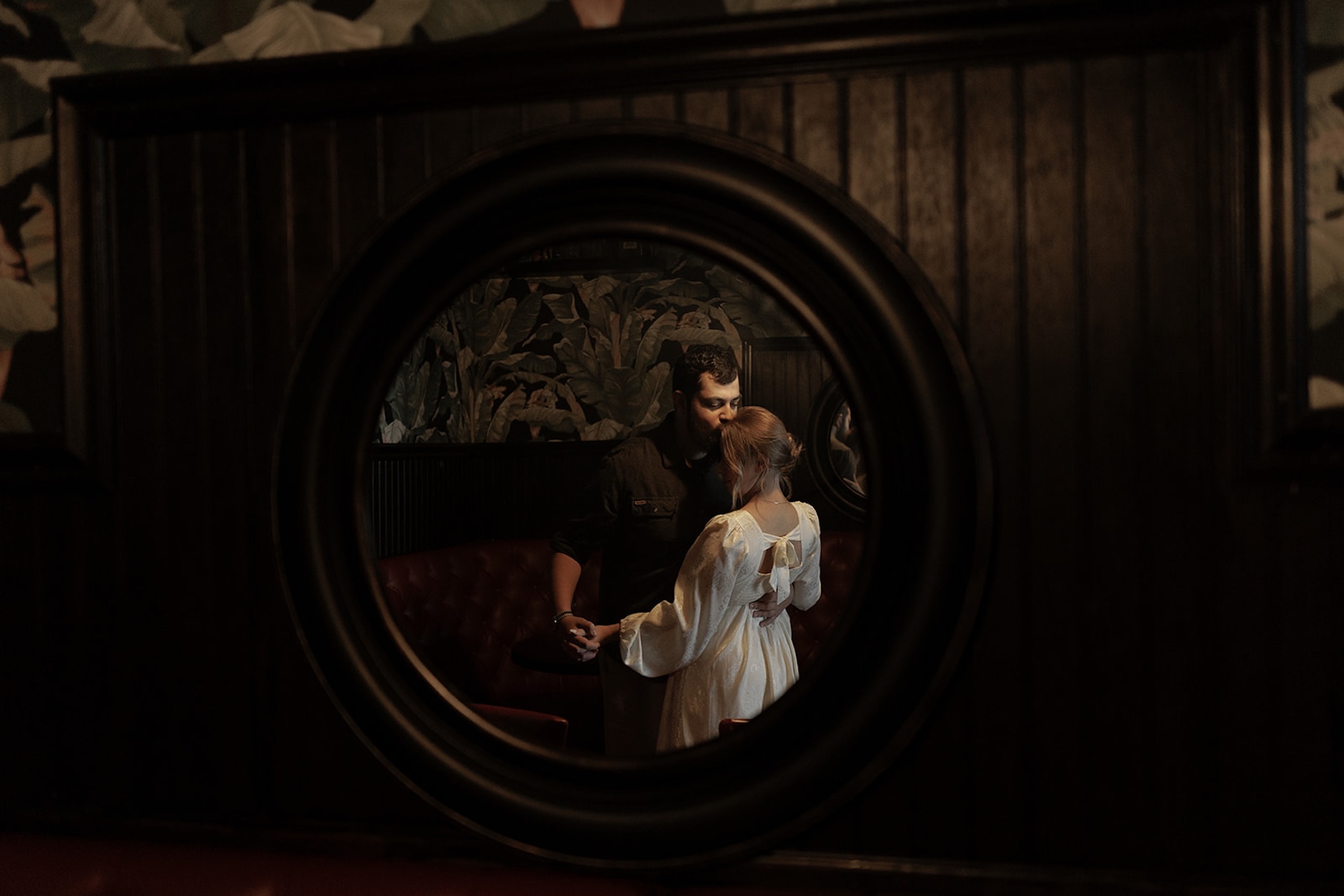 Framed engagement portrait of a couple standing close inside a moody lounge, photographed through architectural details for depth and intimacy.