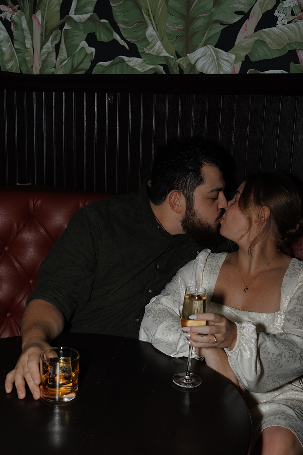 Romantic engagement photo of a couple kissing at a cozy bar booth, holding champagne and whiskey glasses, highlighting moody Engagement Photo Session Ideas with intimate indoor vibes.