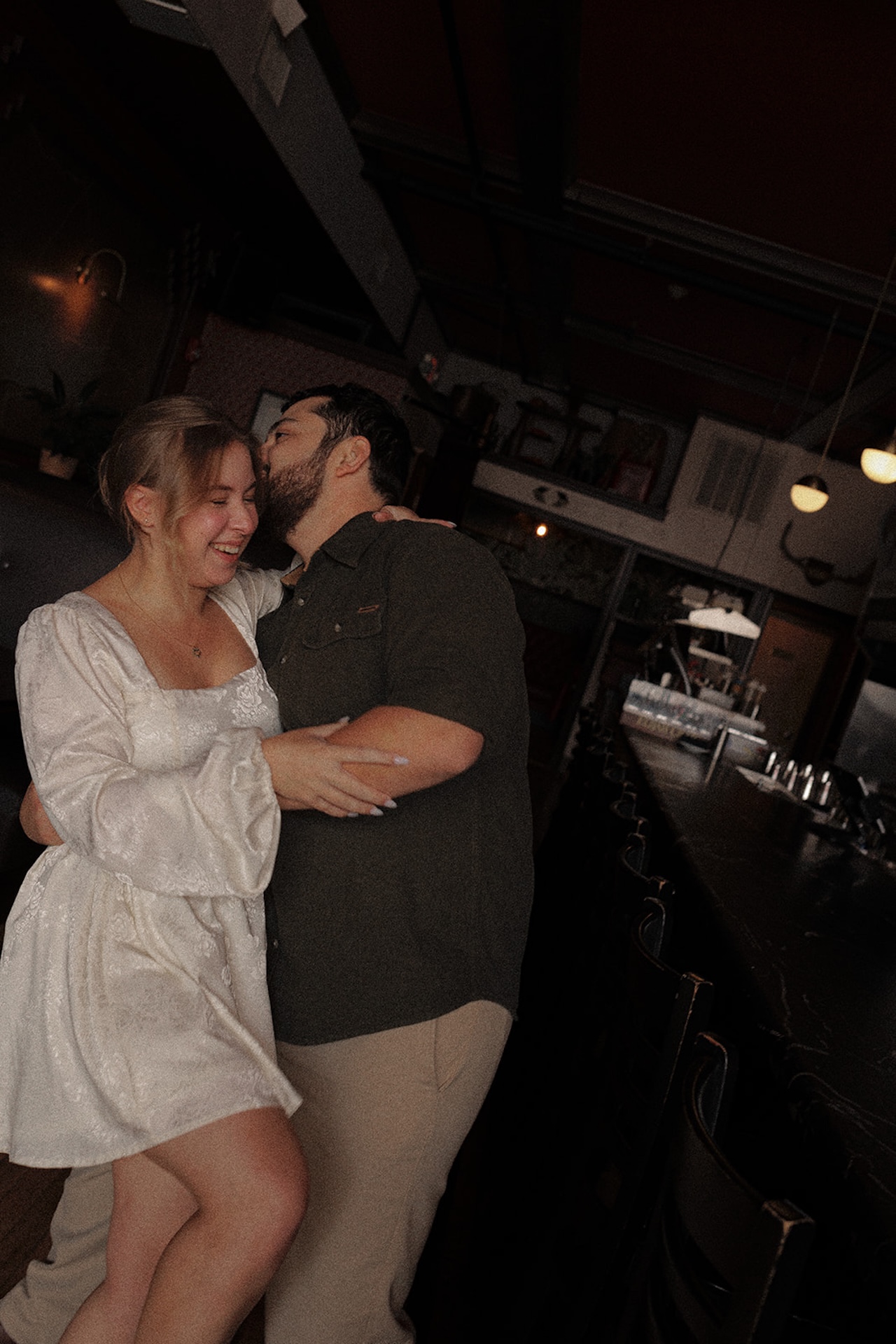 Cozy bar engagement photo of a couple laughing and embracing under warm, moody lighting, capturing intimate Engagement Photo Session Ideas in a stylish indoor setting.
