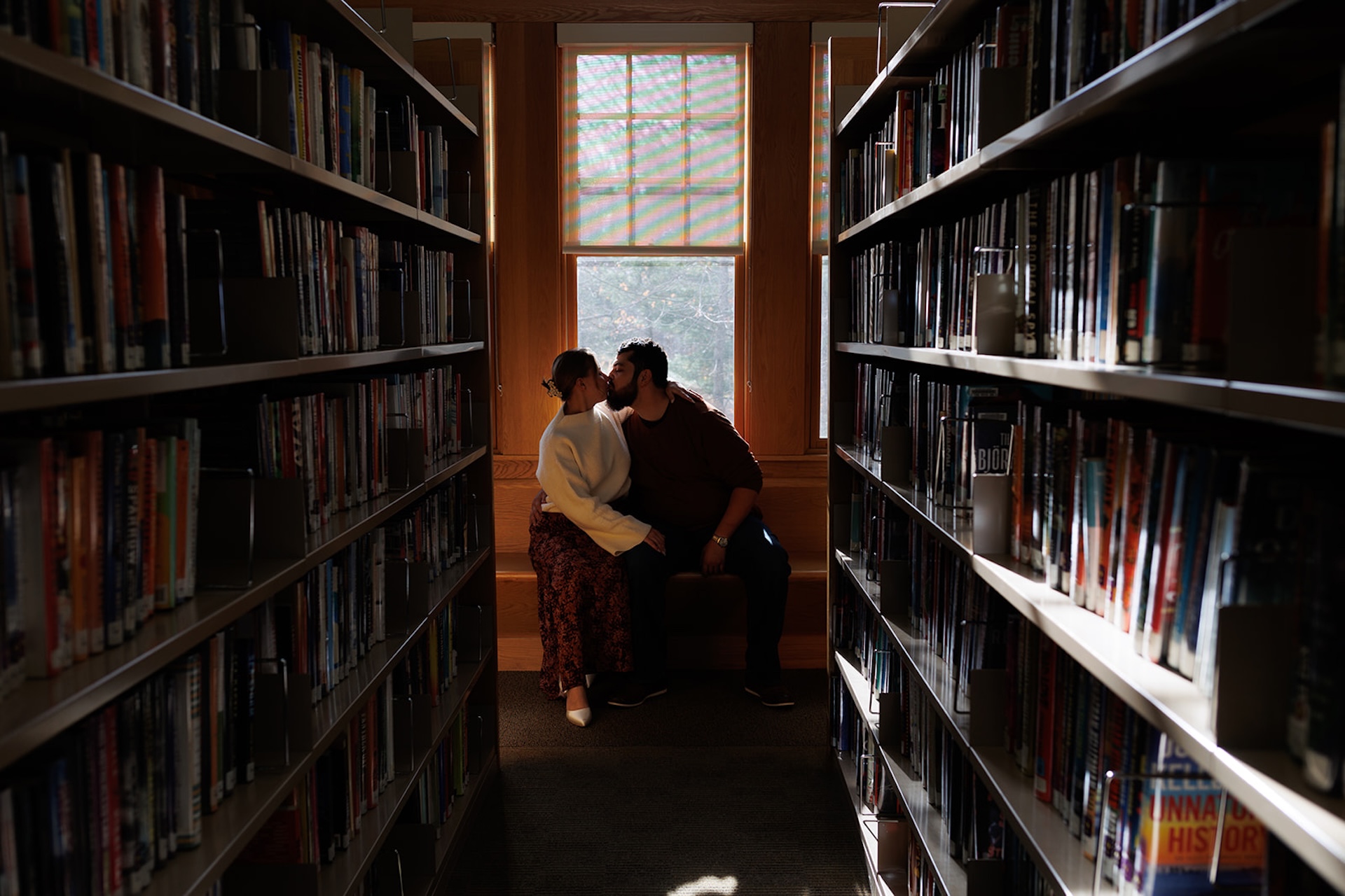 Romantic engagement photo of a couple kissing in a library aisle, softly lit with warm tones and architectural framing.