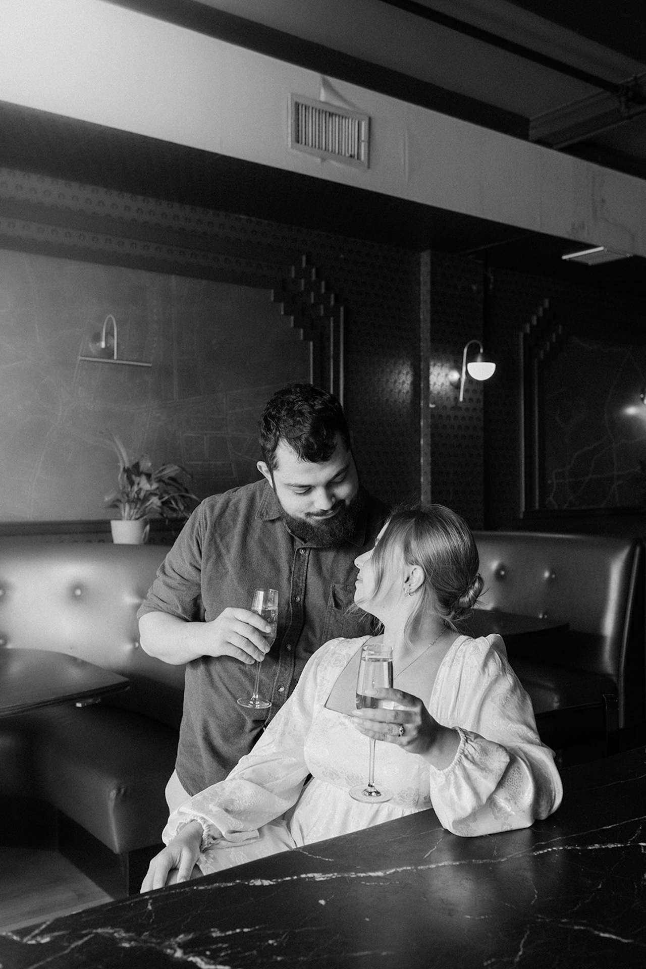 Playful engagement photo of a couple sipping drinks together at a bar, sitting close and laughing in a warm, intimate setting.