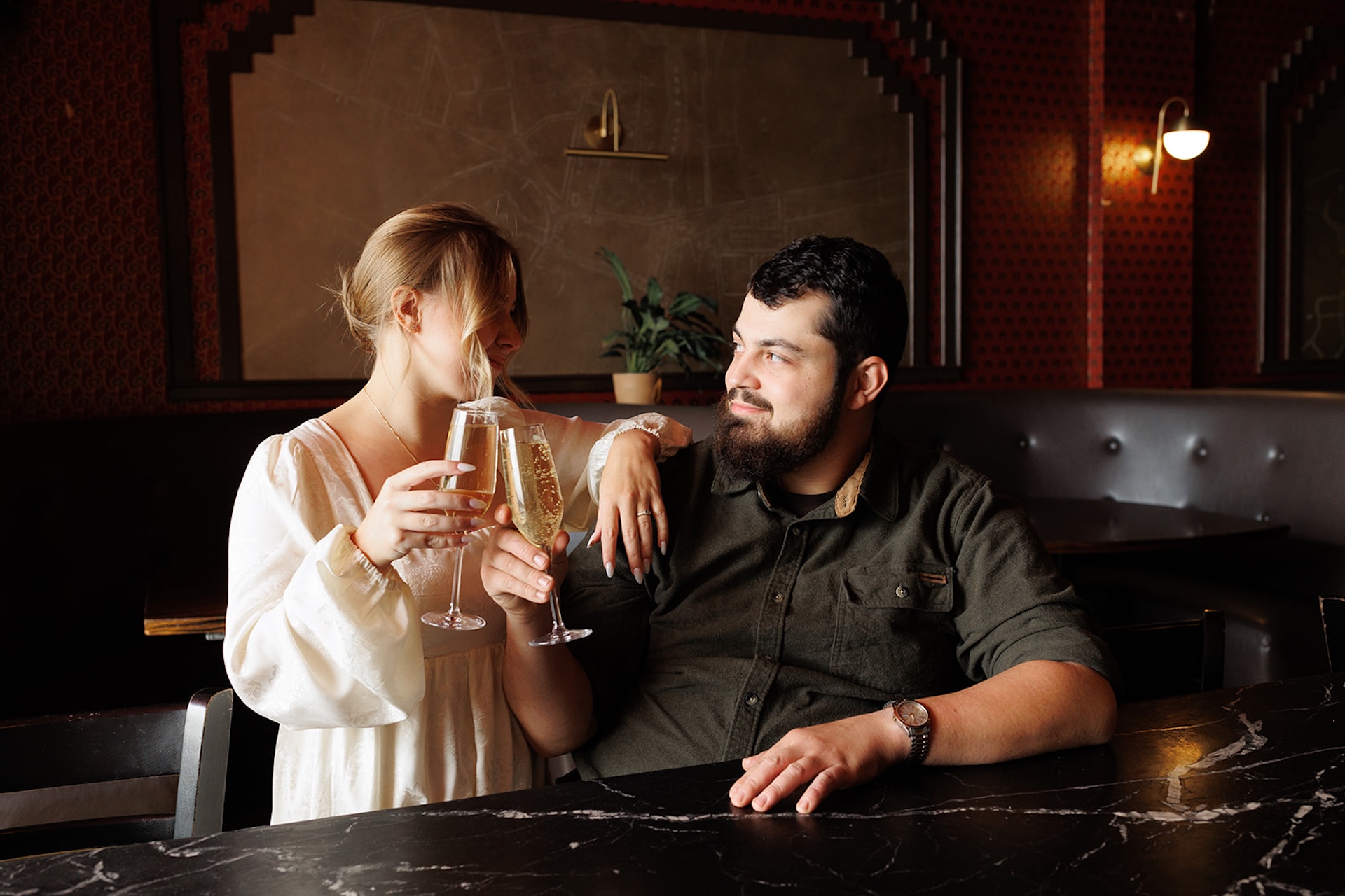 Moody engagement photo of a couple clinking champagne glasses at a bar while smiling at each other, capturing relaxed indoor romance.