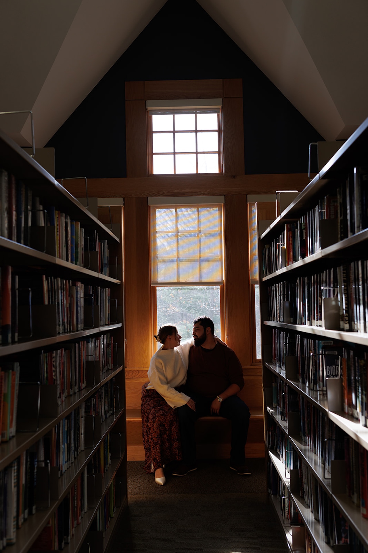Wide engagement photo of a couple seated between tall bookshelves, framed symmetrically by rows of books and natural window light.