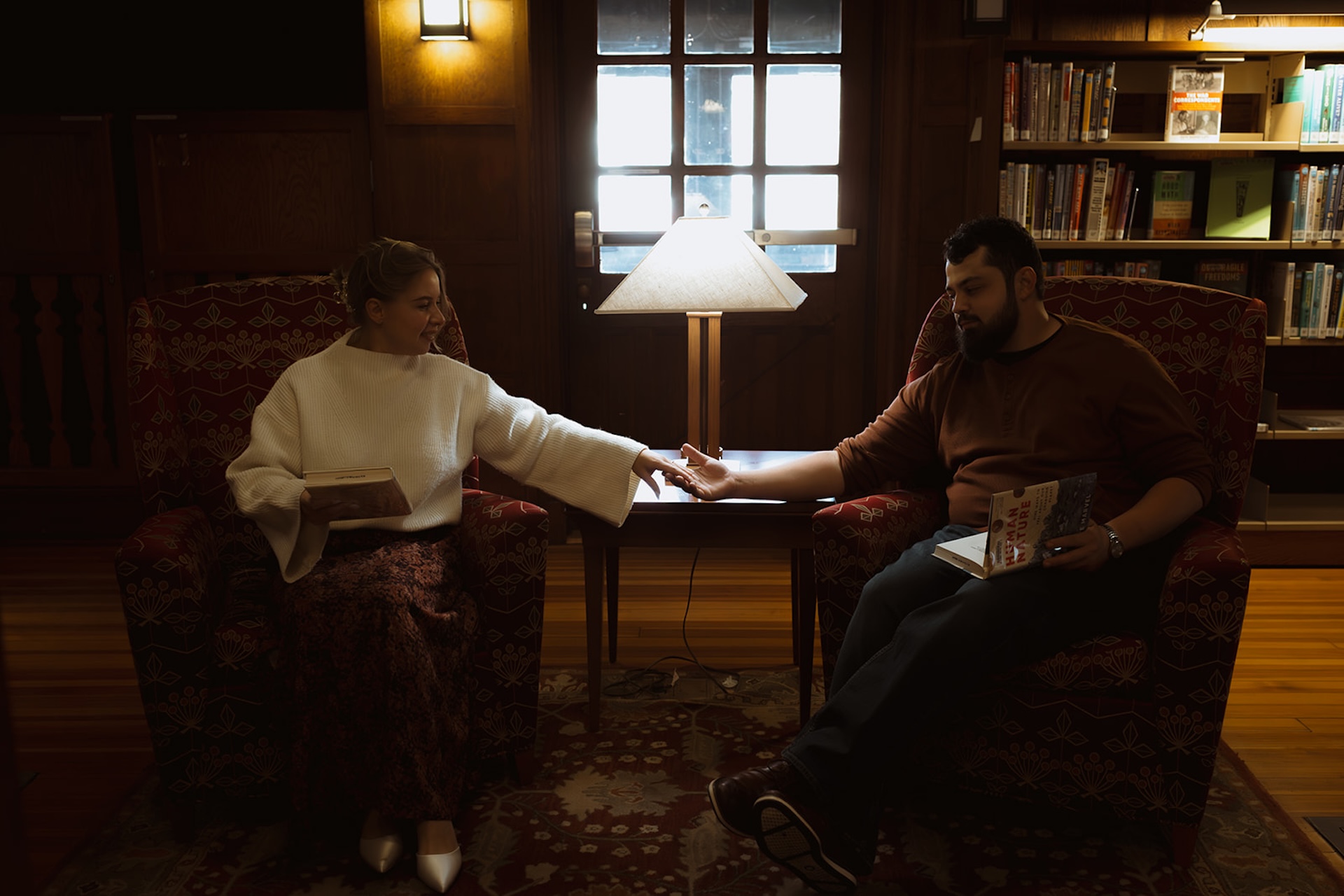 Intimate engagement photo of a couple seated across from each other in a library lounge, holding hands and reading books under soft, warm lighting.
