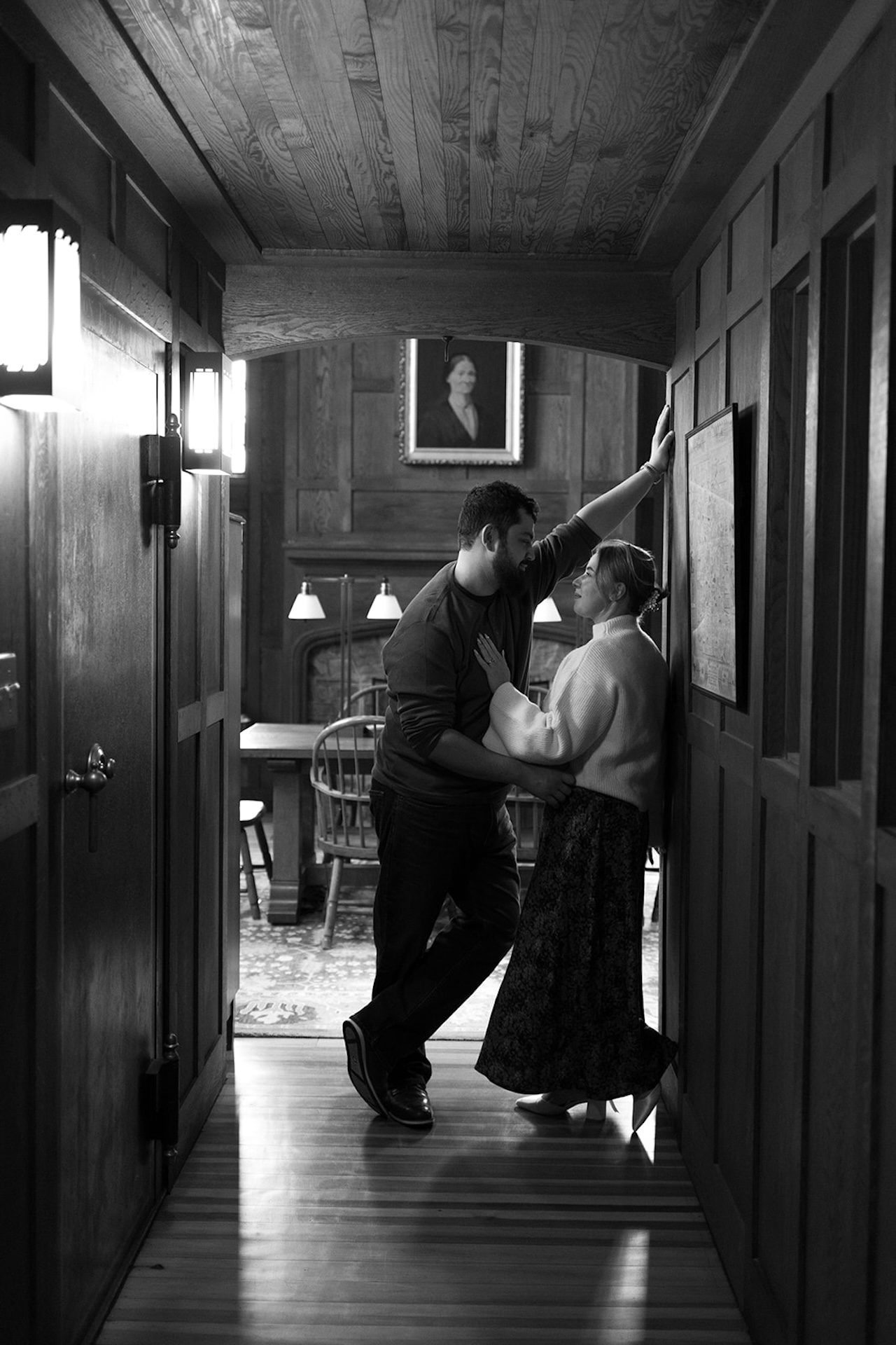 Black and white engagement photo of a couple leaning into each other in a narrow hallway, sharing a relaxed and candid moment indoors.
