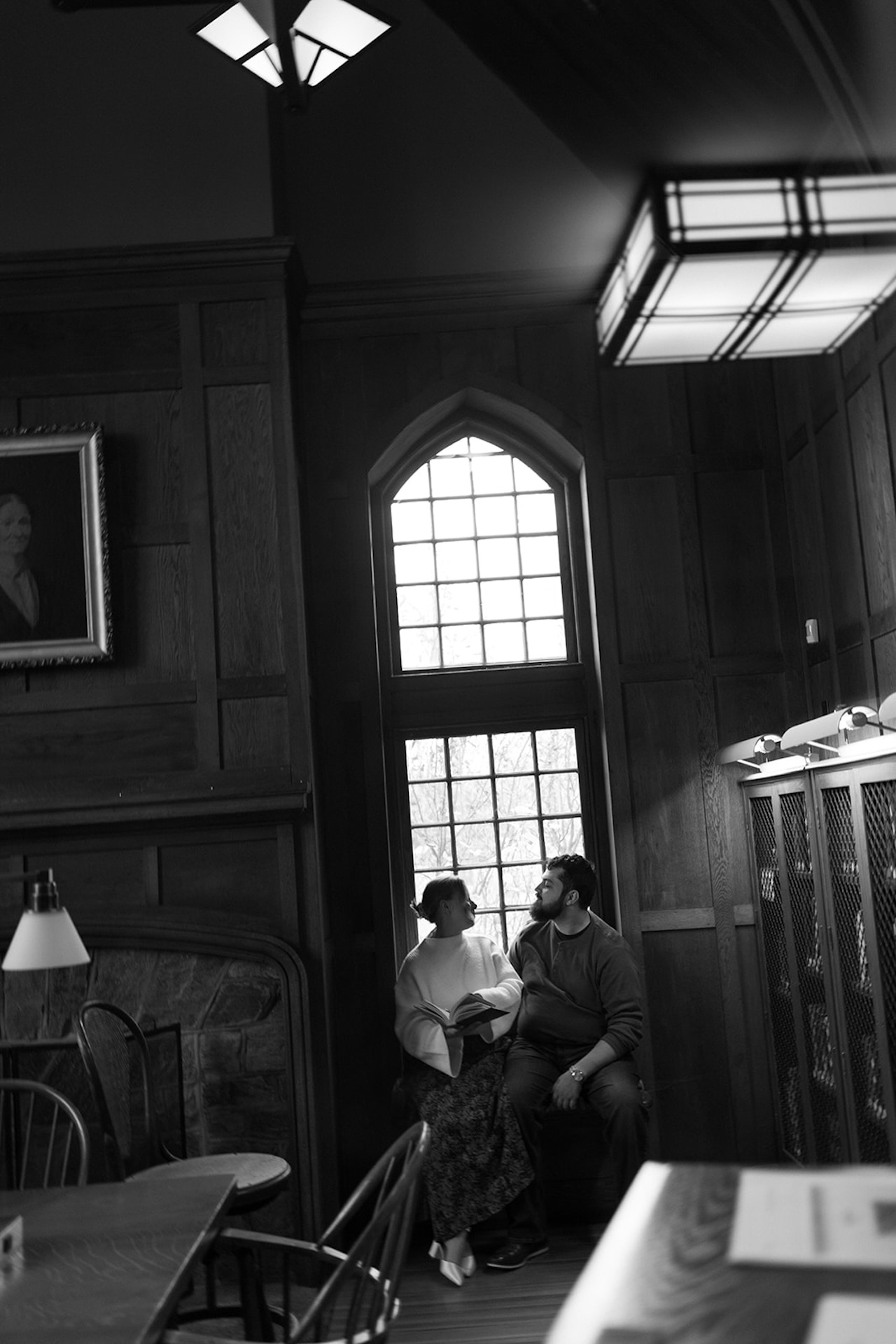 Black and white engagement photo of a couple sitting together beneath tall arched windows in a historic library, surrounded by wood-paneled walls.
