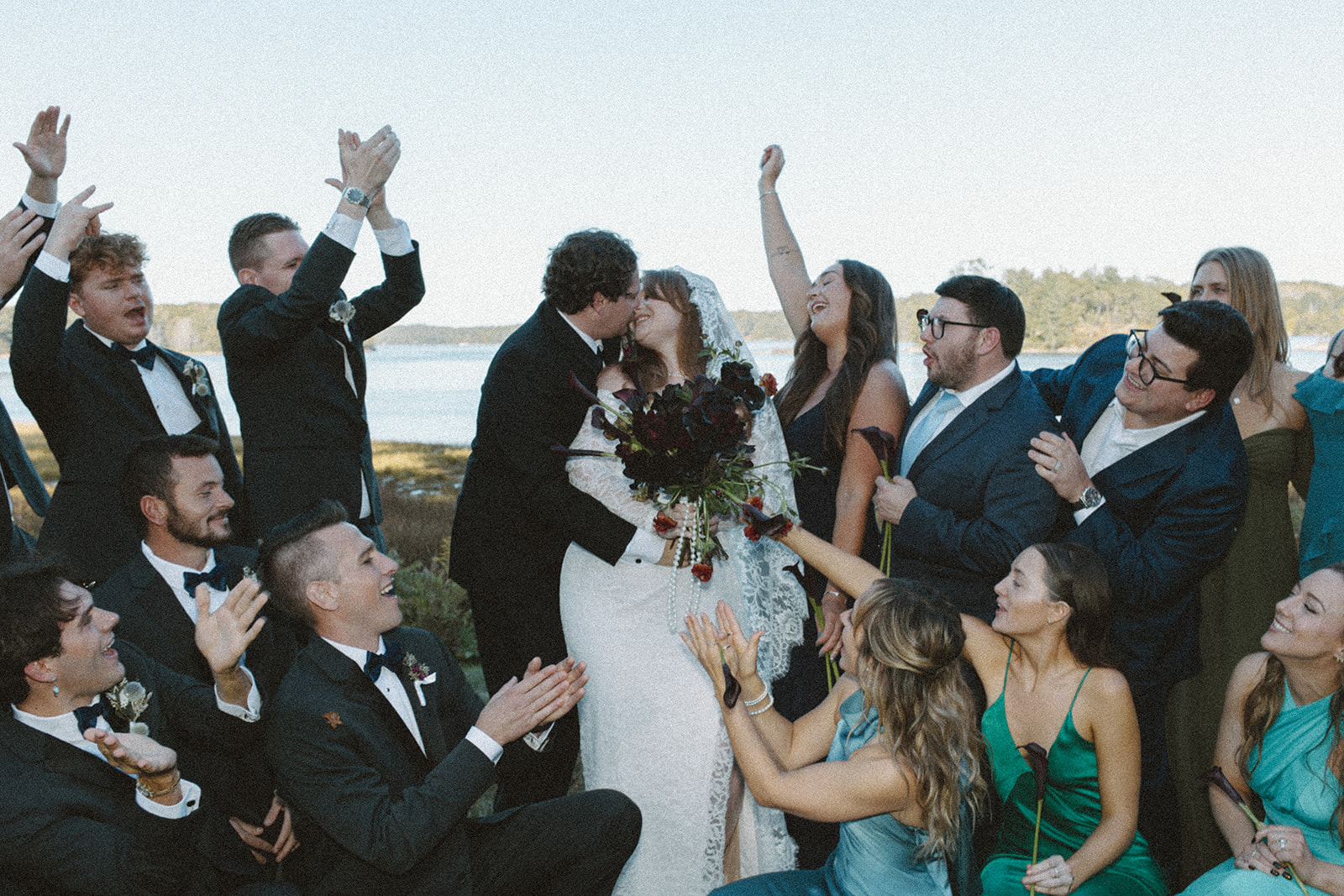 A film wedding party photo of the bride and groom leaning in for a kiss as their bridesmaids and groomsmen celebrate around them.