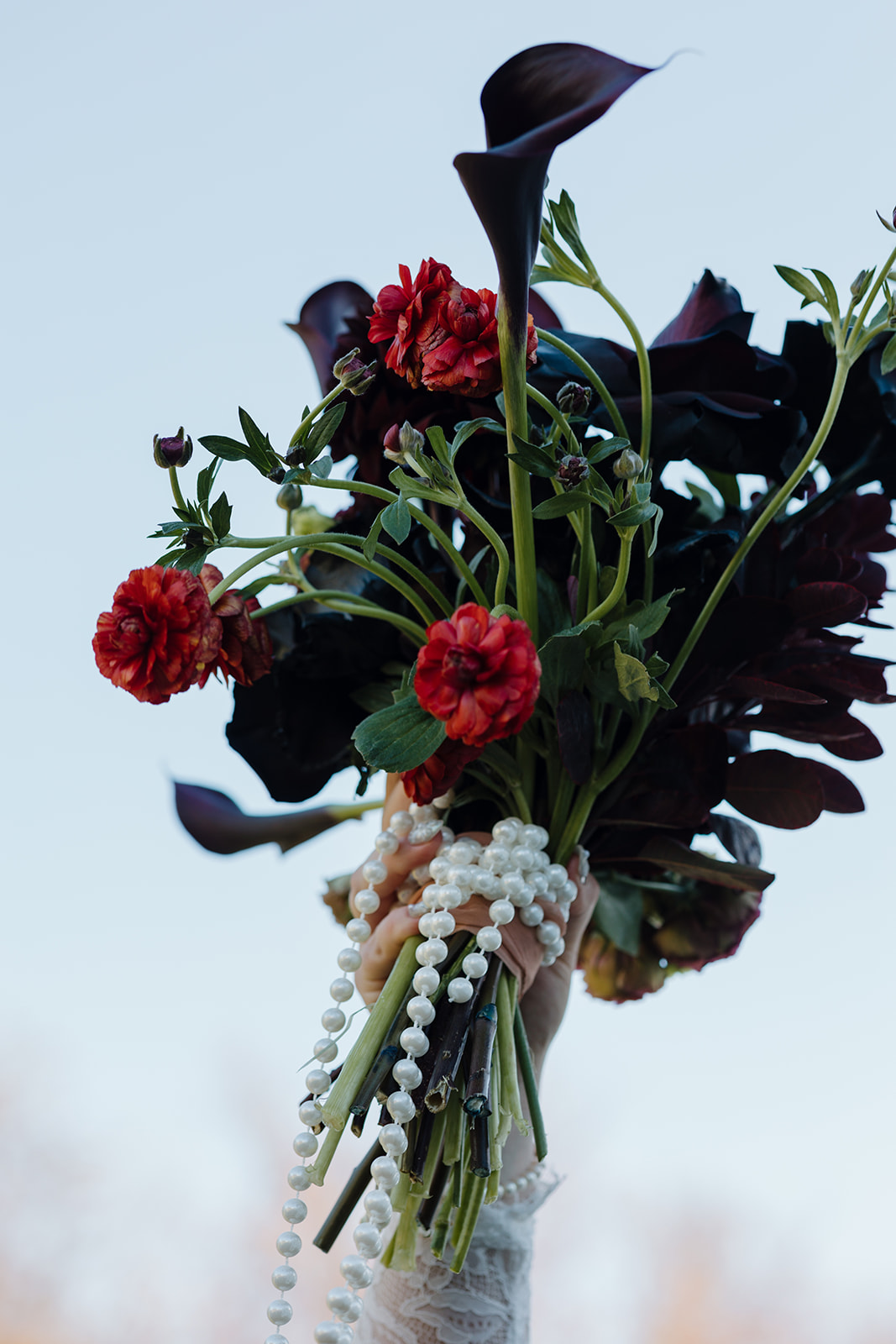 A detail photo of the bride holding her moody wedding bouquet in the sky with dark maroon, orange, and red florals and strings of pearls during her fall destination wedding in Maine